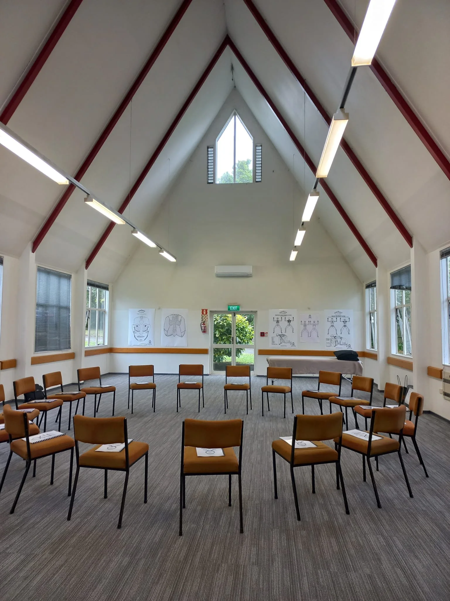 A meeting room with chairs arranged in a circle, marked with paper and symbols on each chair, with a high ceiling, windows on all walls, and diagrams of human anatomy on the walls.