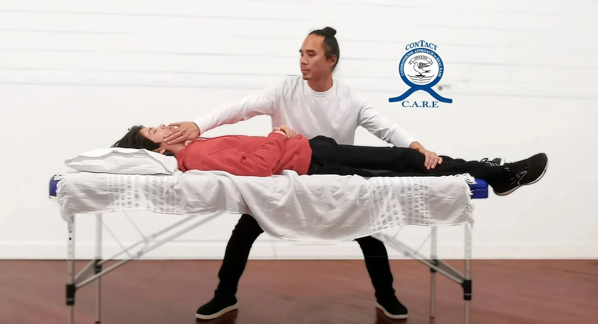 A man performing CPR on a woman lying on a hospital bed in a training room. The man is kneeling beside the bed, with one hand on the woman's chin and the other hand on her forehead to open her airway. The woman is wearing a red jacket, and the man is dressed in a white shirt and black pants. There is a logo on the wall that says 'CONTACT C.A.R.E' in the background.