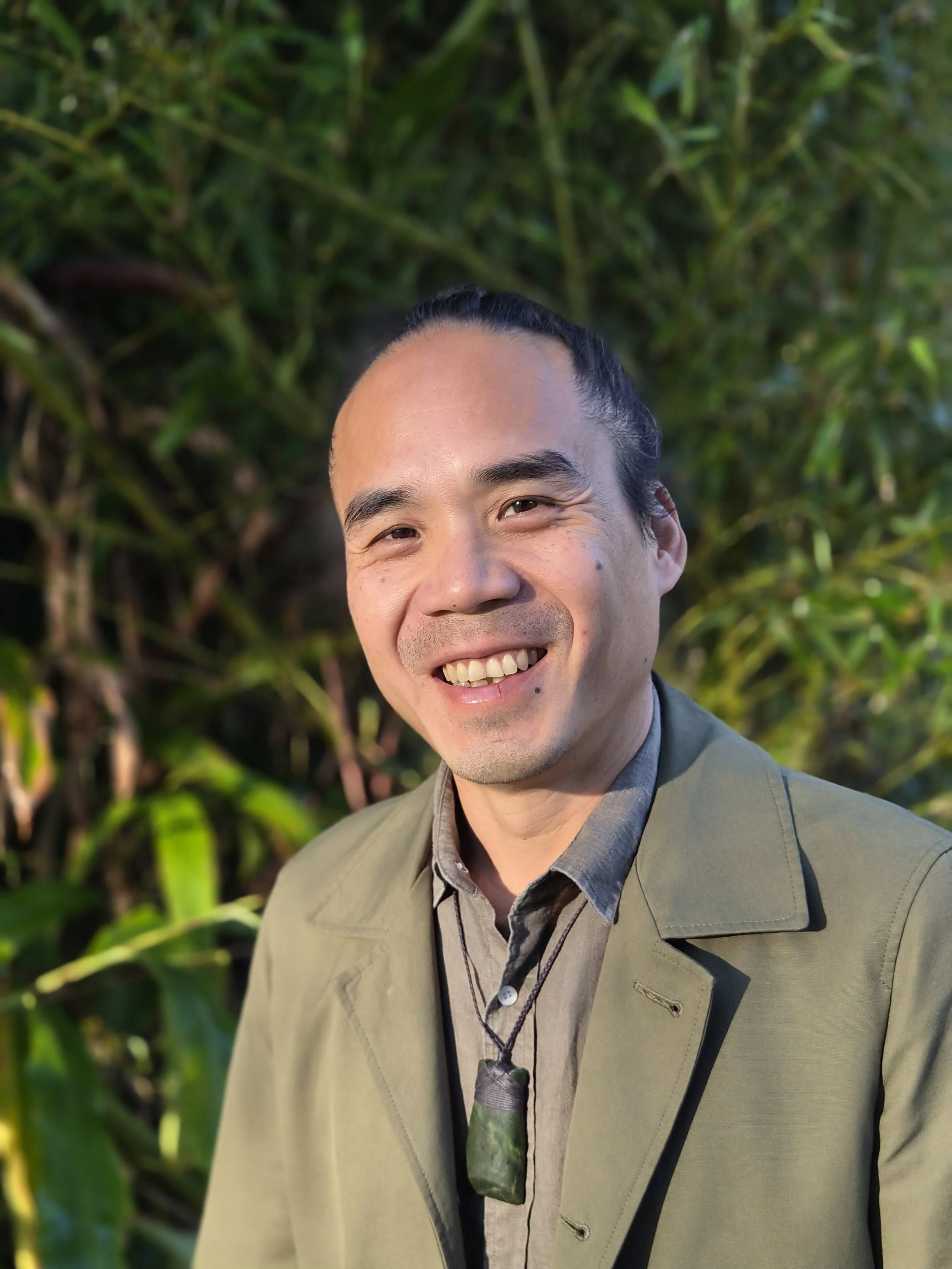 A smiling man in a suit and shirt standing outdoors in front of green foliage.