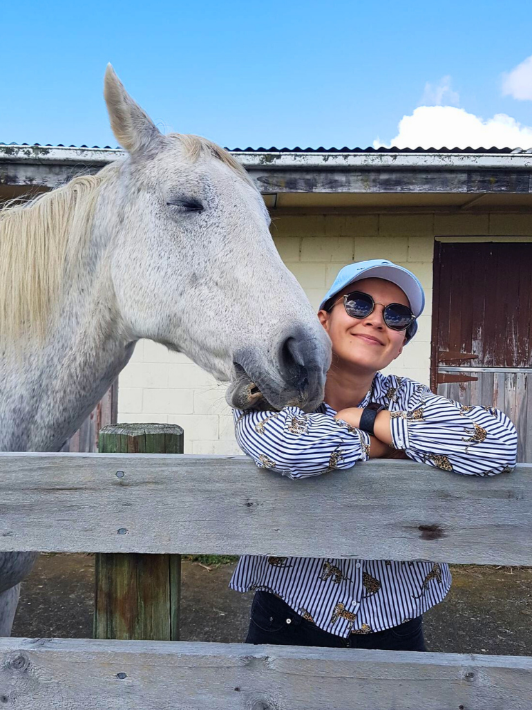 A woman wearing sunglasses, a light blue cap, and a striped shirt with leopard print, leaning on a wooden fence with her arms crossed, smiling as a white horse nuzzles her face outdoors near a building with a cloudy blue sky overhead.