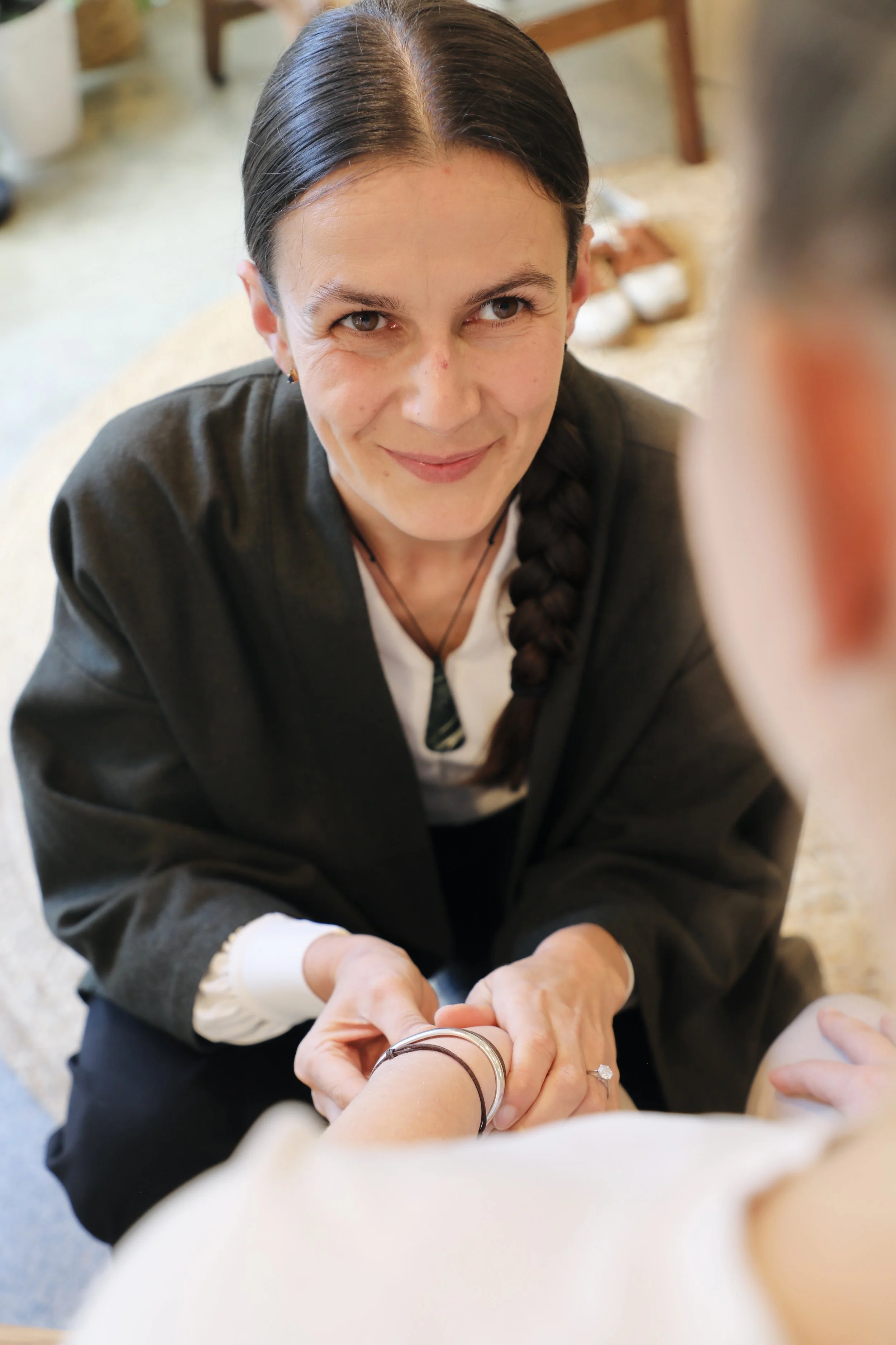A woman with dark hair in a braid is smiling while holding hands with another person in the foreground. The woman is wearing a dark jacket and is indoors, with furniture and shoes visible in the background.