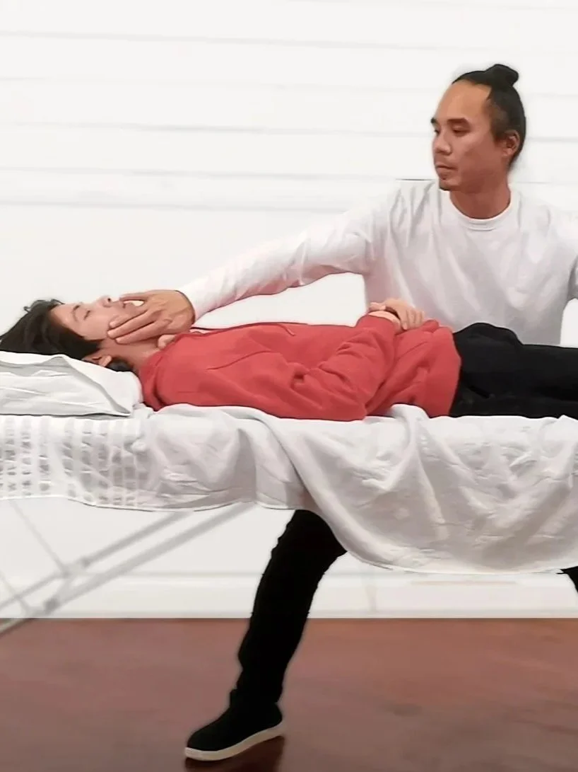 A man with long hair tied in a bun holding a woman in a red sweater on a bed. The woman is lying down with her eyes closed, and the man is gently holding her face and hand. The bed has white sheets and a pillow, and the background features a plain white wall.
