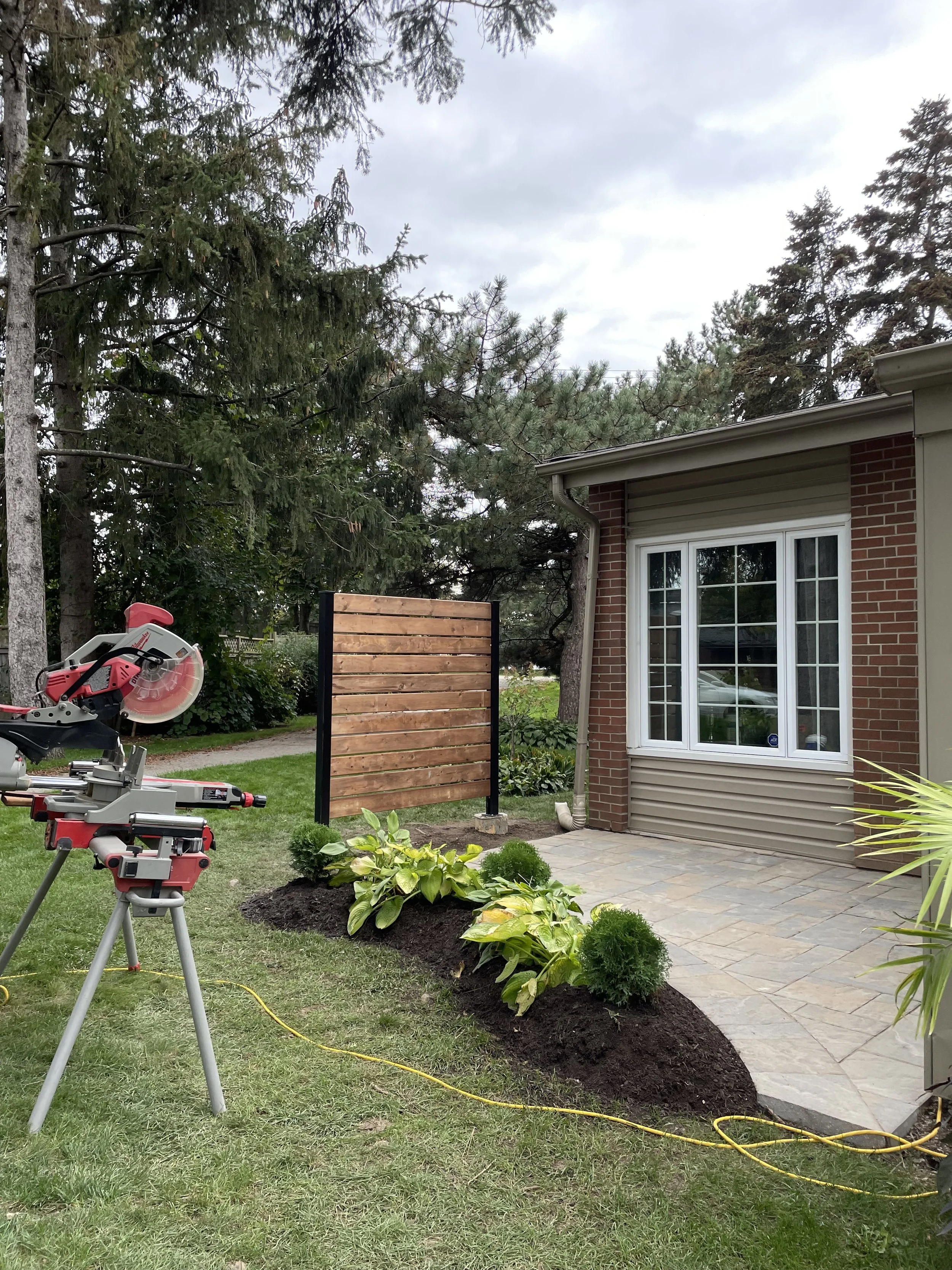 A backyard with a new garden bed of plants and flowers in front of a house, a miter saw on a stand, a wooden privacy screen, and trees in the background.