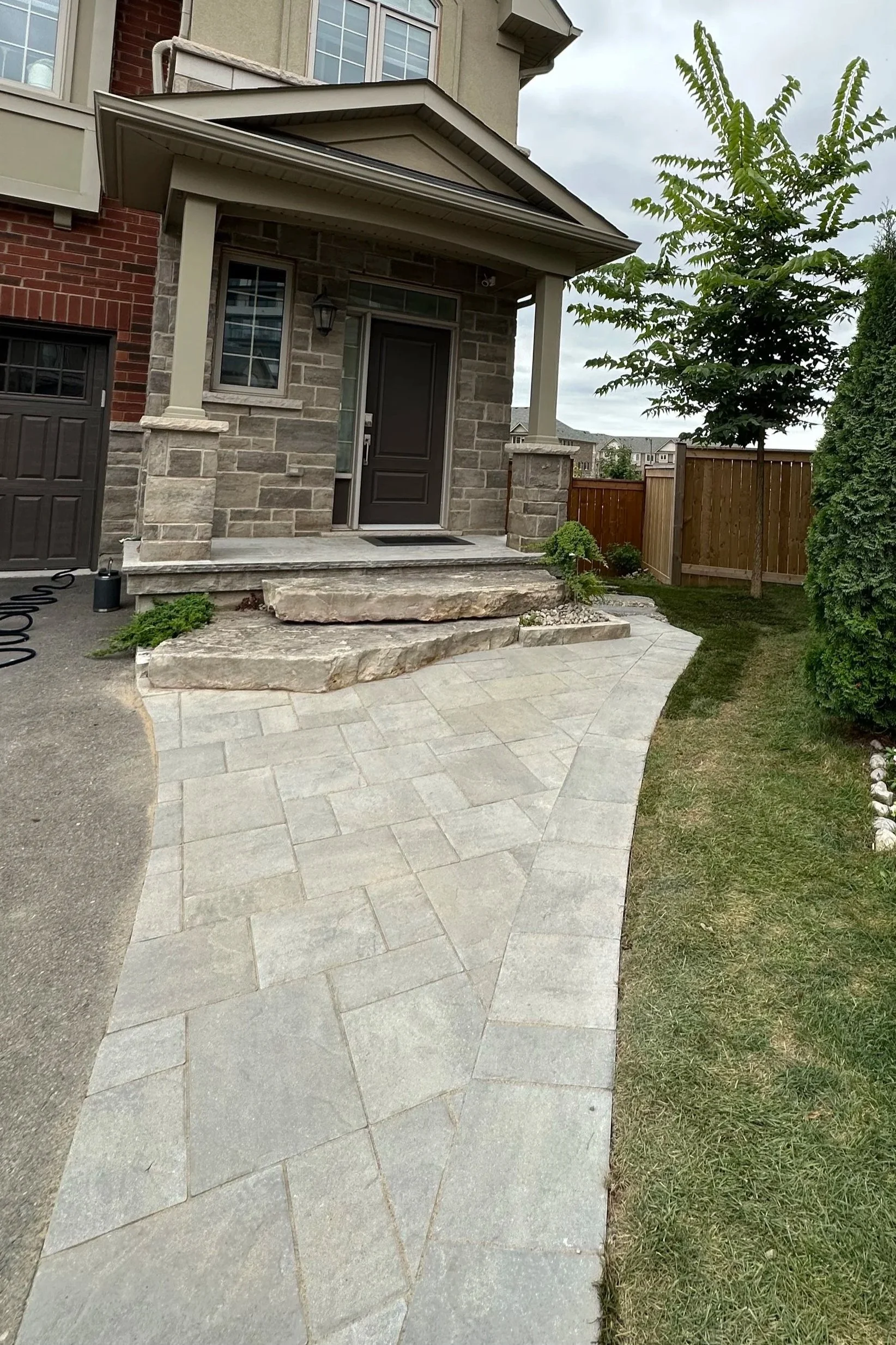 Newly paved stone pathway leading to the front porch of a house, with a small garden and a wooden fence on the side.