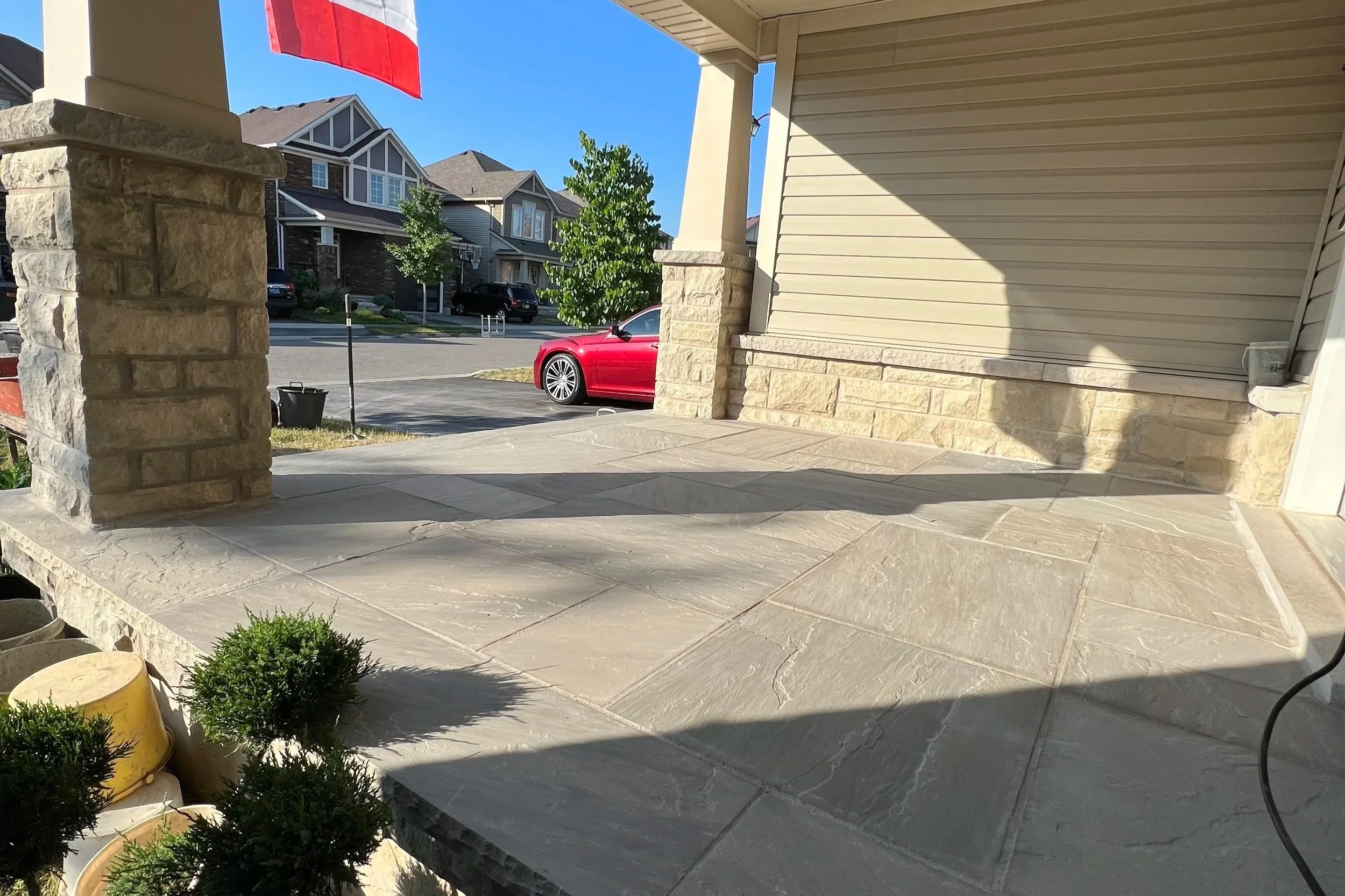 Front porch with stone columns and beige siding, featuring a shadow of a hanging flower pot and plants in pots near the entrance. A red car is parked on the street, with a neighborhood of modern houses in the background under a clear blue sky.
