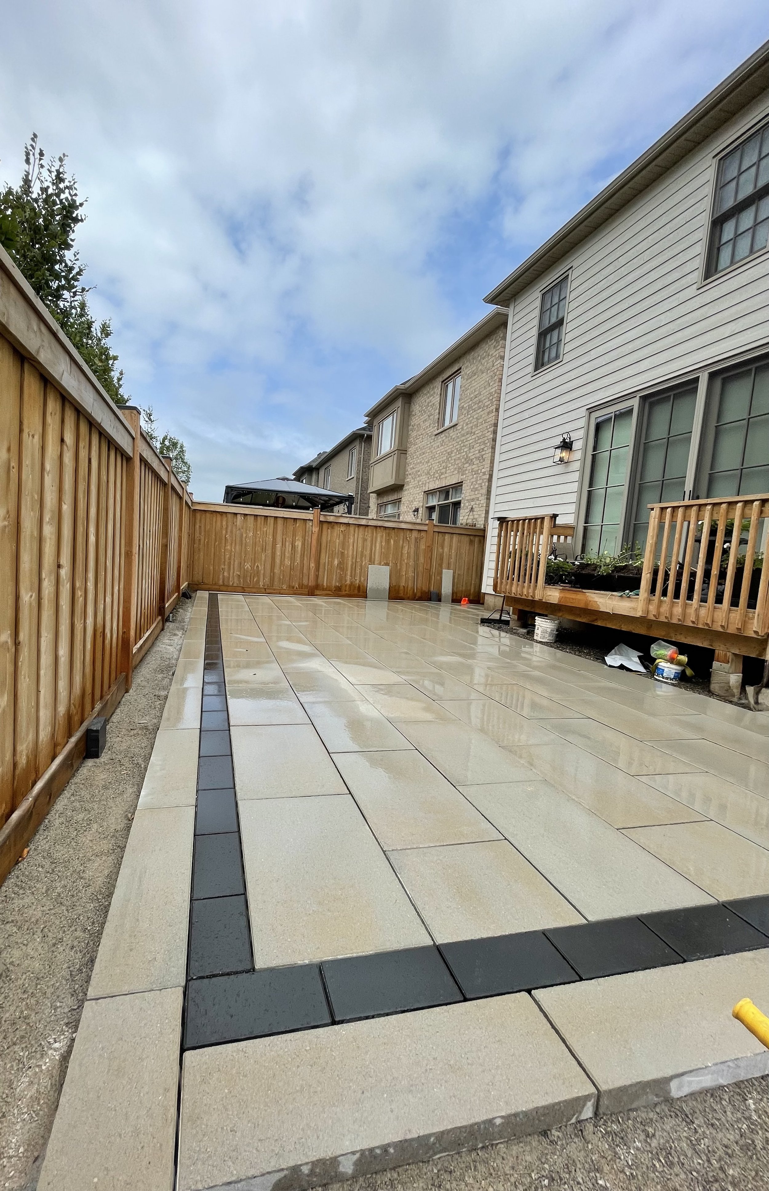 Finished patio with beige and black tiles, wooden fence on the left, and the side of a house with large windows on the right.