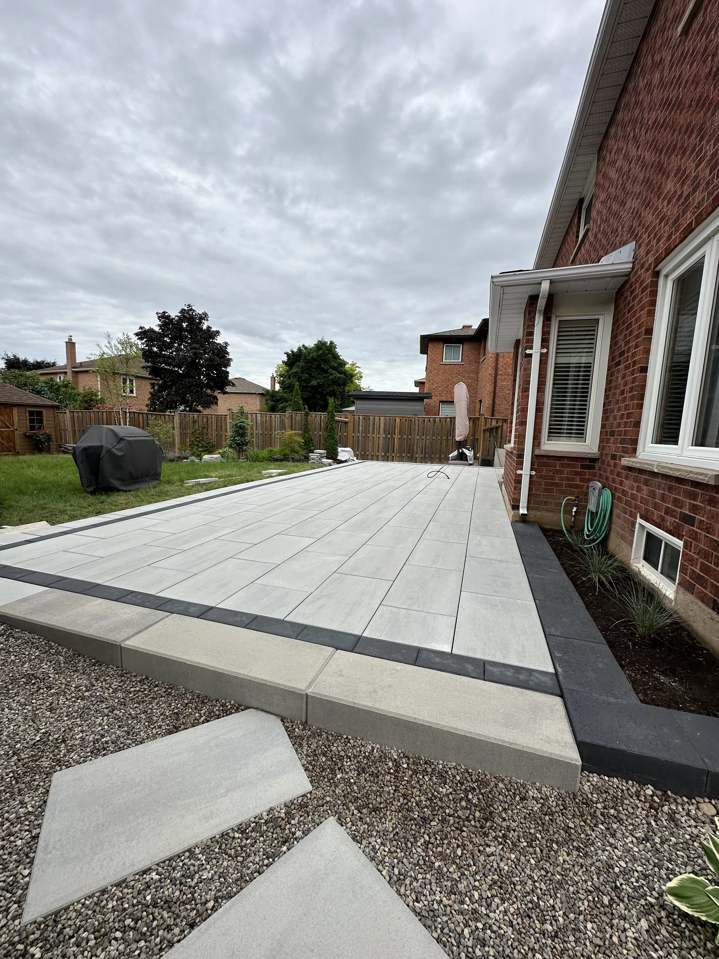 A newly paved backyard patio with large white tiles bordered by black bricks, attached to a red brick house with white window frames. There is a garden hose on the side of the house, a covered grill on the grass, and a wooden fence enclosing the yard