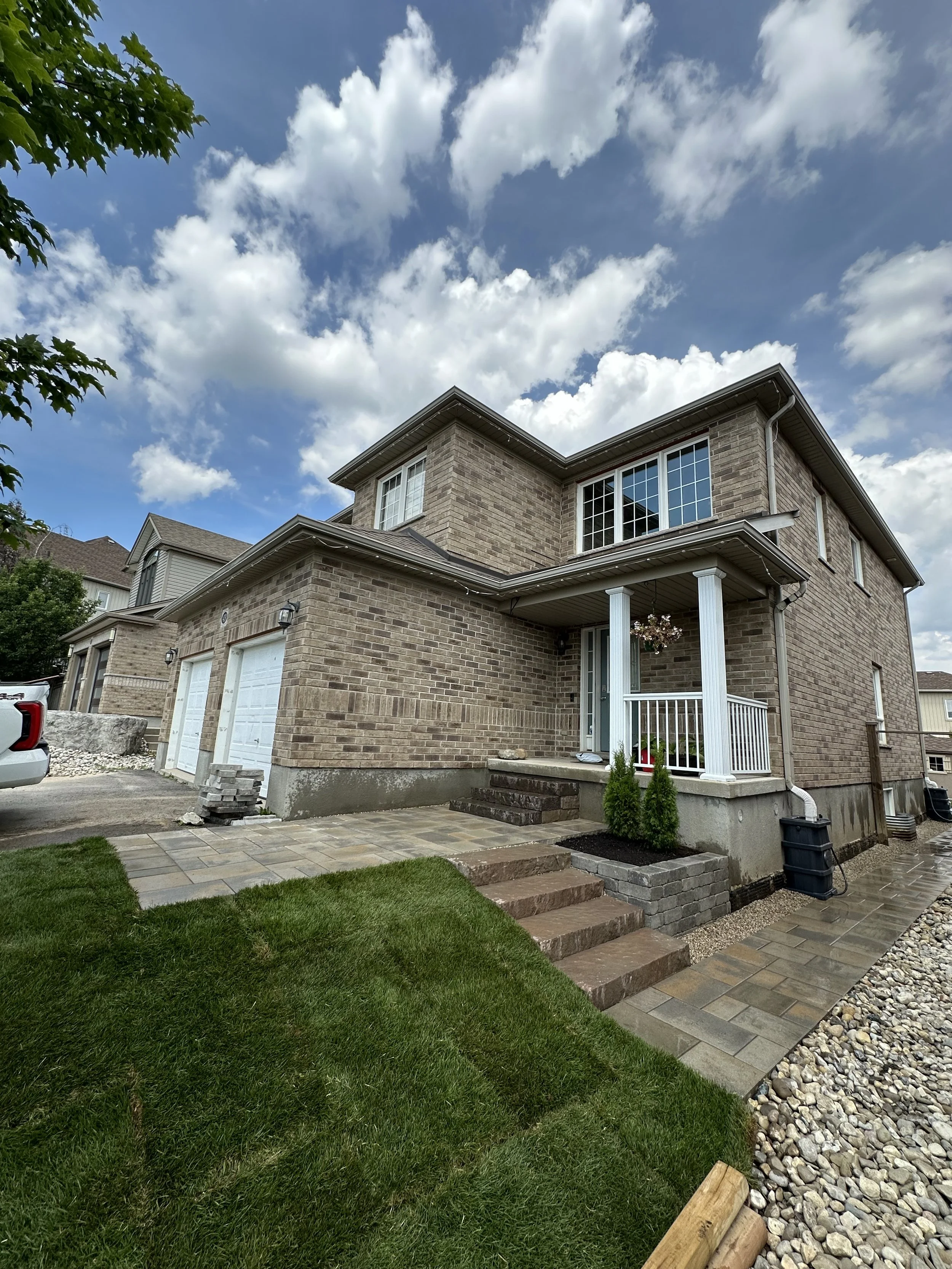Two-story brick house with a front porch, white columns, and stairs leading to a door. The yard has green grass, pavers, and decorative stones. Overcast sky with scattered clouds.