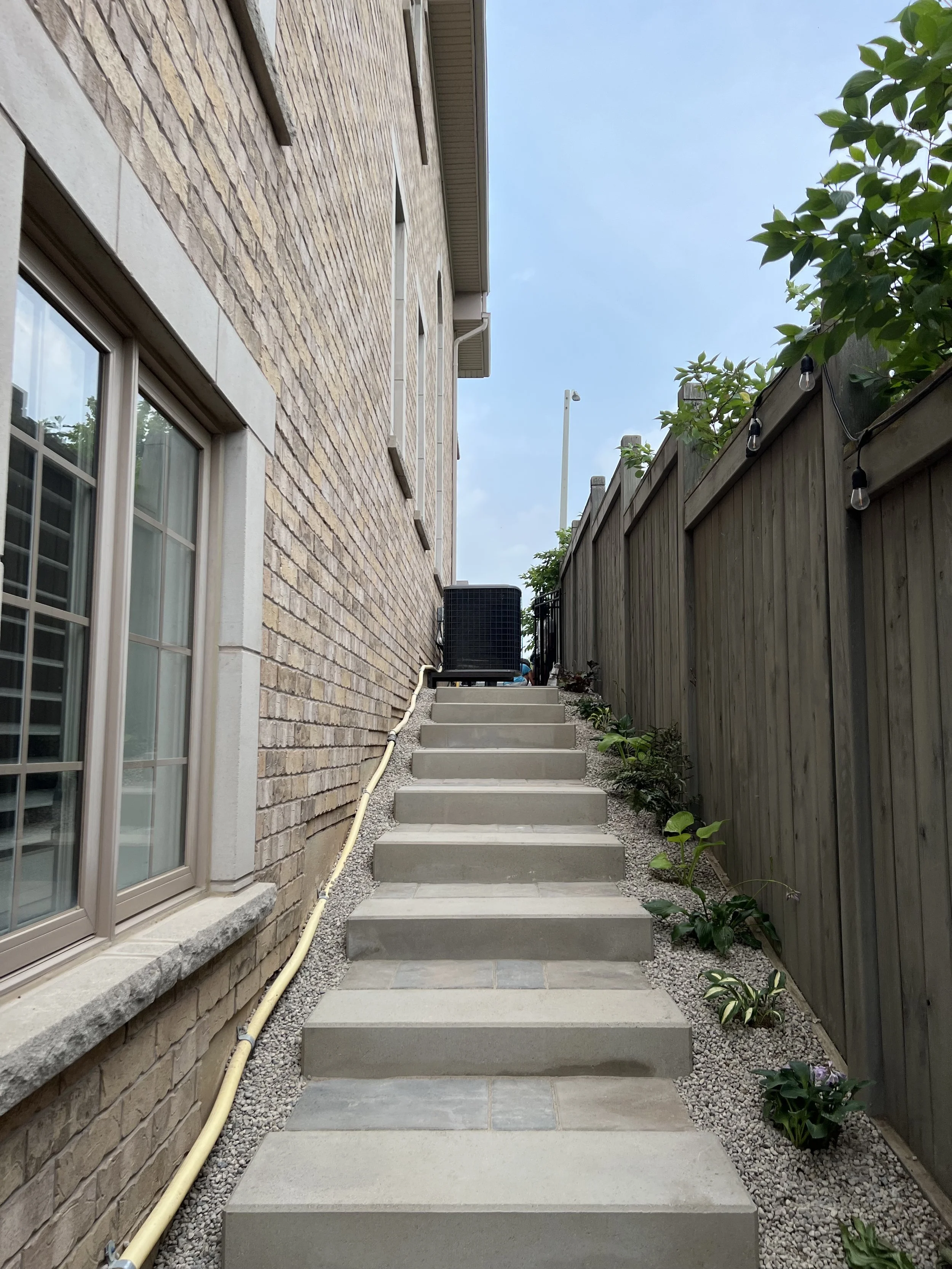 Image of outdoor concrete stairs leading uphill alongside a brick house on the left and a wooden fence on the right, with small plants growing along the fence and string lights hanging.