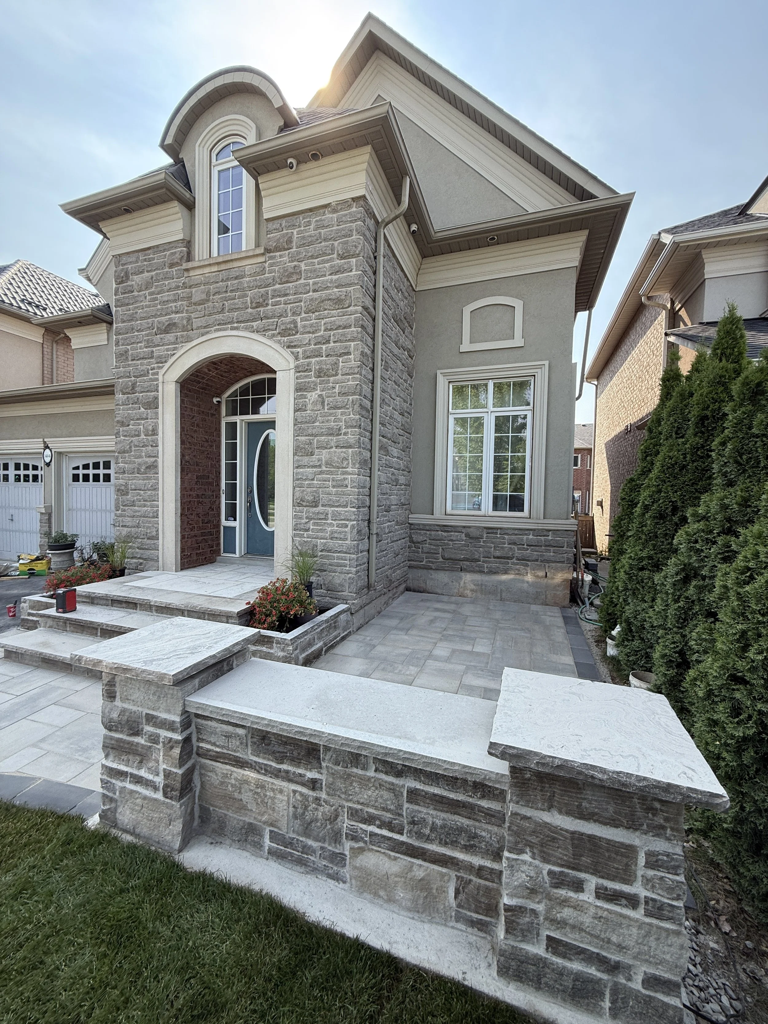 Front view of a house with stone and stucco exterior, arched entryway, large window, and a small porch with steps, surrounded by a stone wall and landscaping.