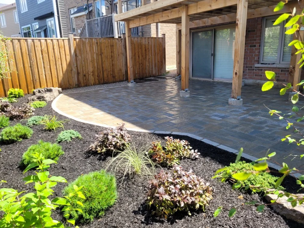 A backyard patio area with a curved stone or brick paved surface, partially covered by a wooden pergola with vertical support beams. The patio is adjacent to a brick house with sliding glass doors. There is a landscaped garden bed with various small 