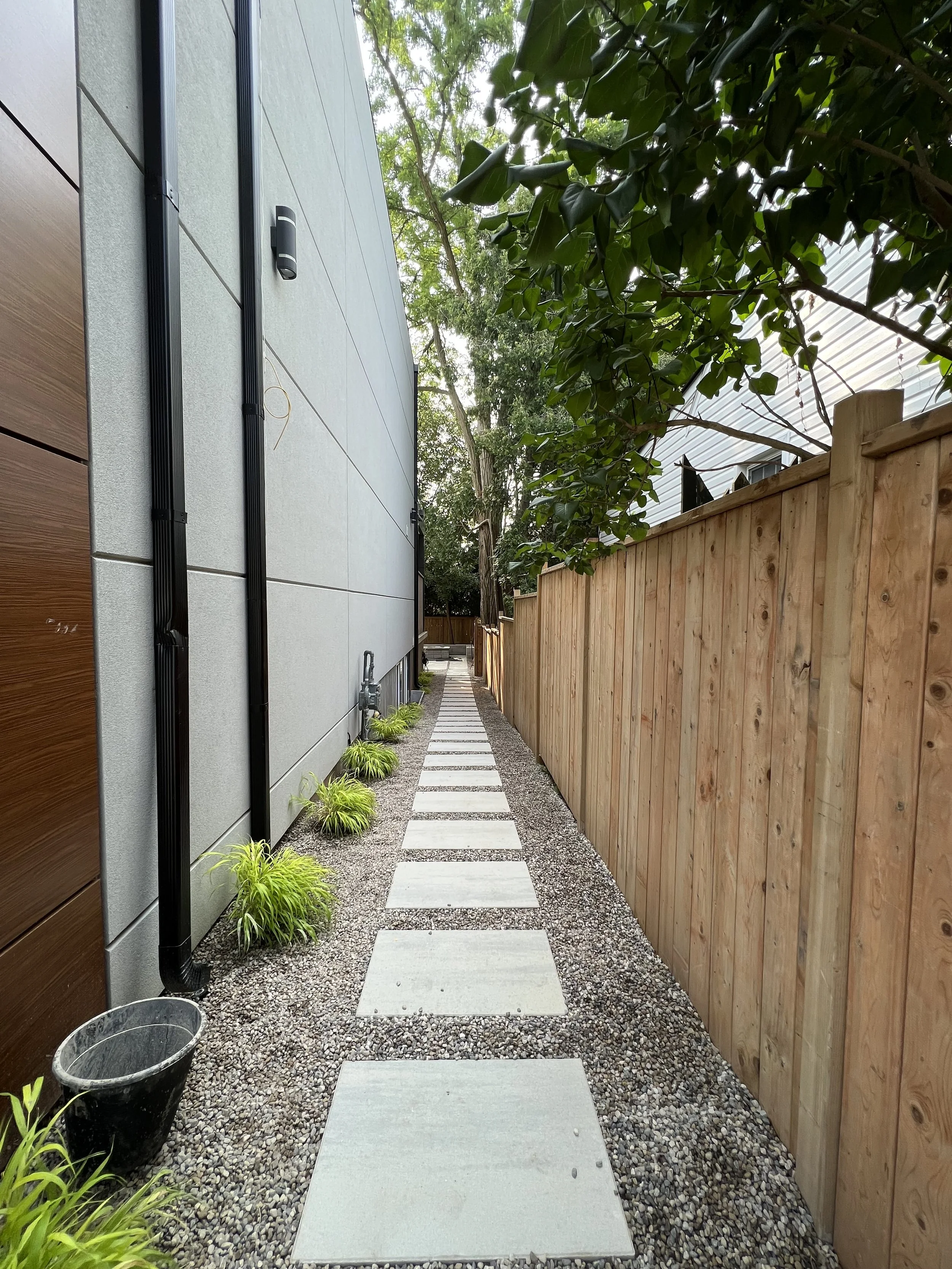 A narrow outdoor pathway with square concrete stepping stones on a gravel surface, bordered by a modern house wall on the left and a wooden fence on the right, with trees and greenery overhead.