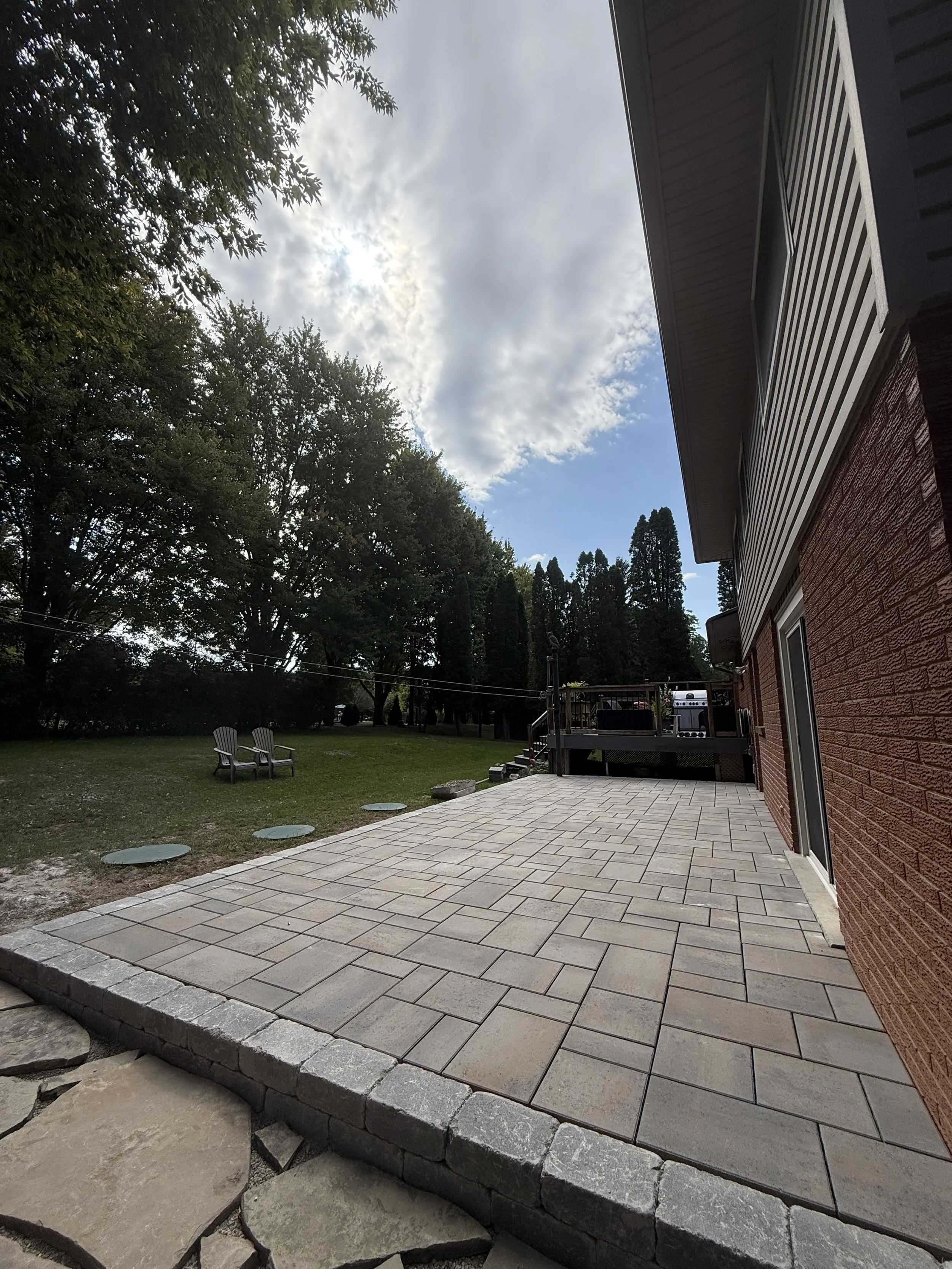 Backyard with stone-paved patio, two Adirondack chairs on grass, large trees in the background, partly cloudy sky, house with siding and brick exterior.