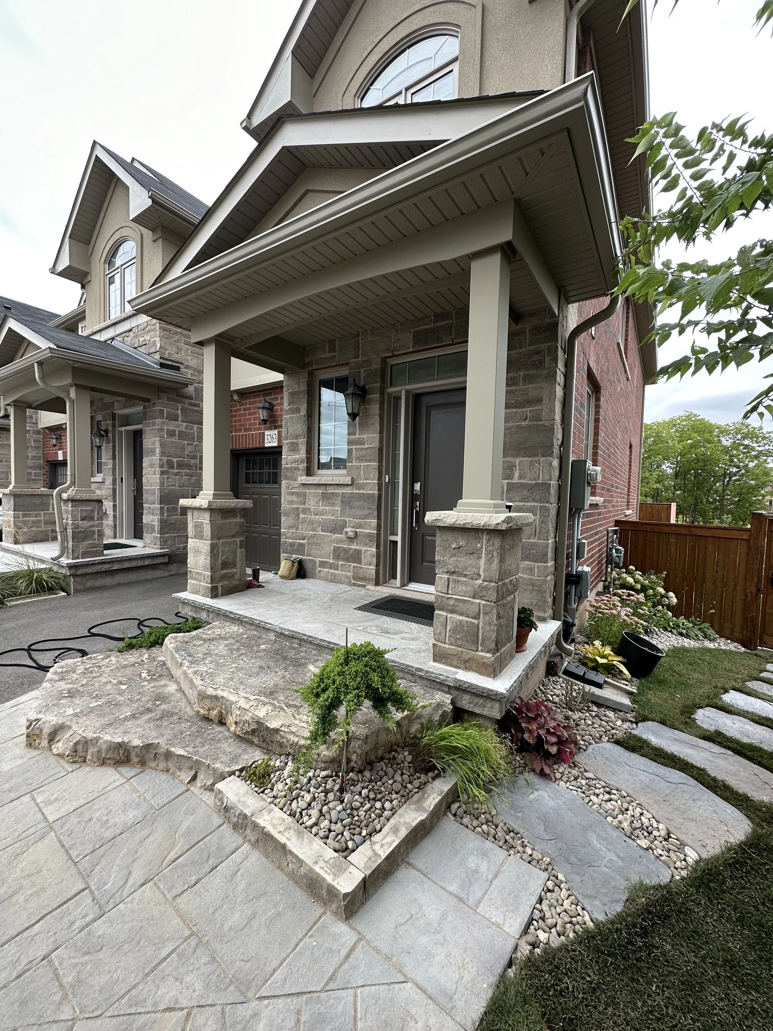 Front yard of a modern house with a stone porch, steps, and landscaping including rocks, small shrubs, and a flower bed. The house has a brick and stone exterior with a covered entryway and a black front door.
