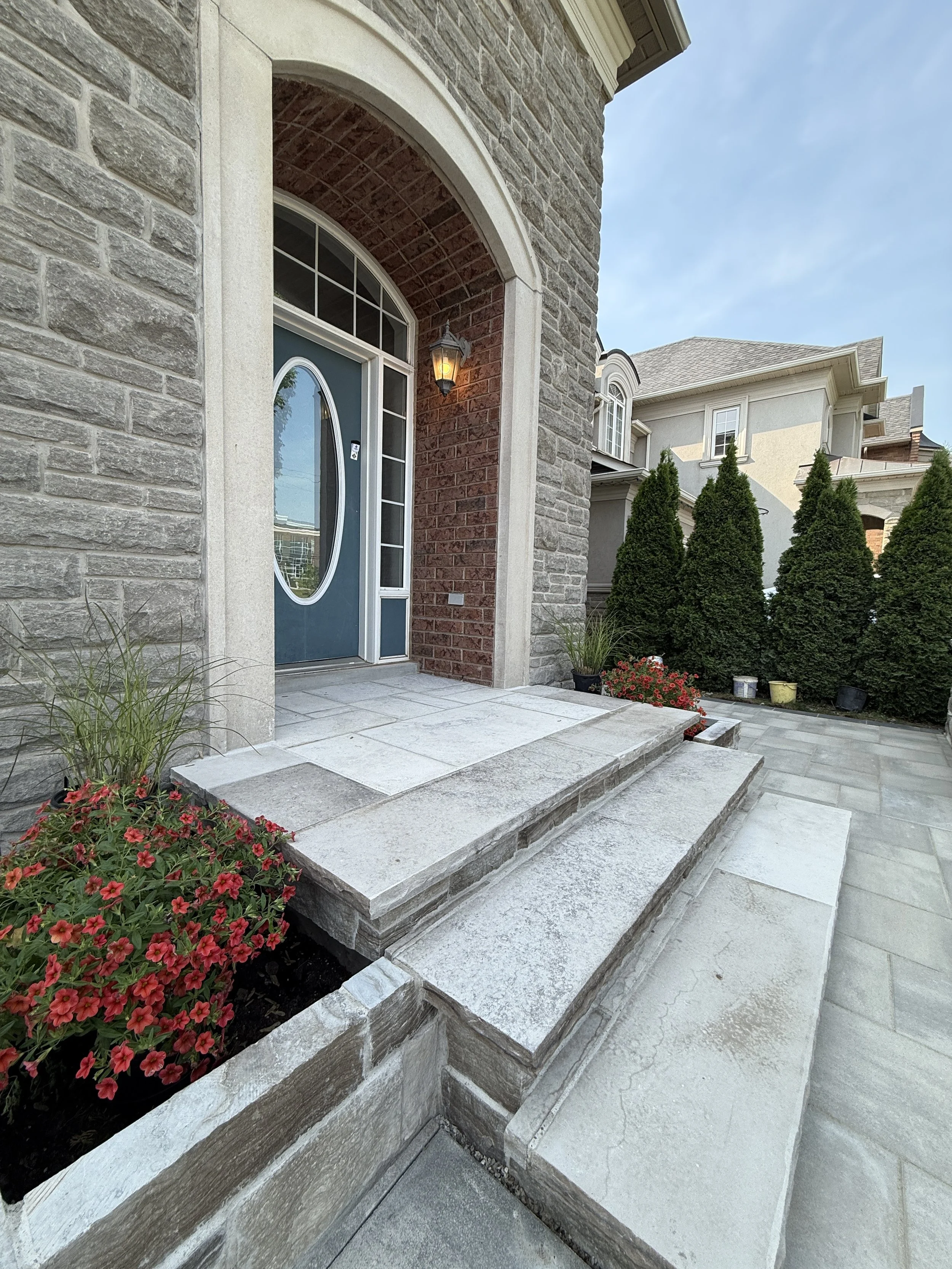 Front porch with stone steps, potted plants with red flowers, green shrubs, and a house with brick and siding exterior under a blue sky.