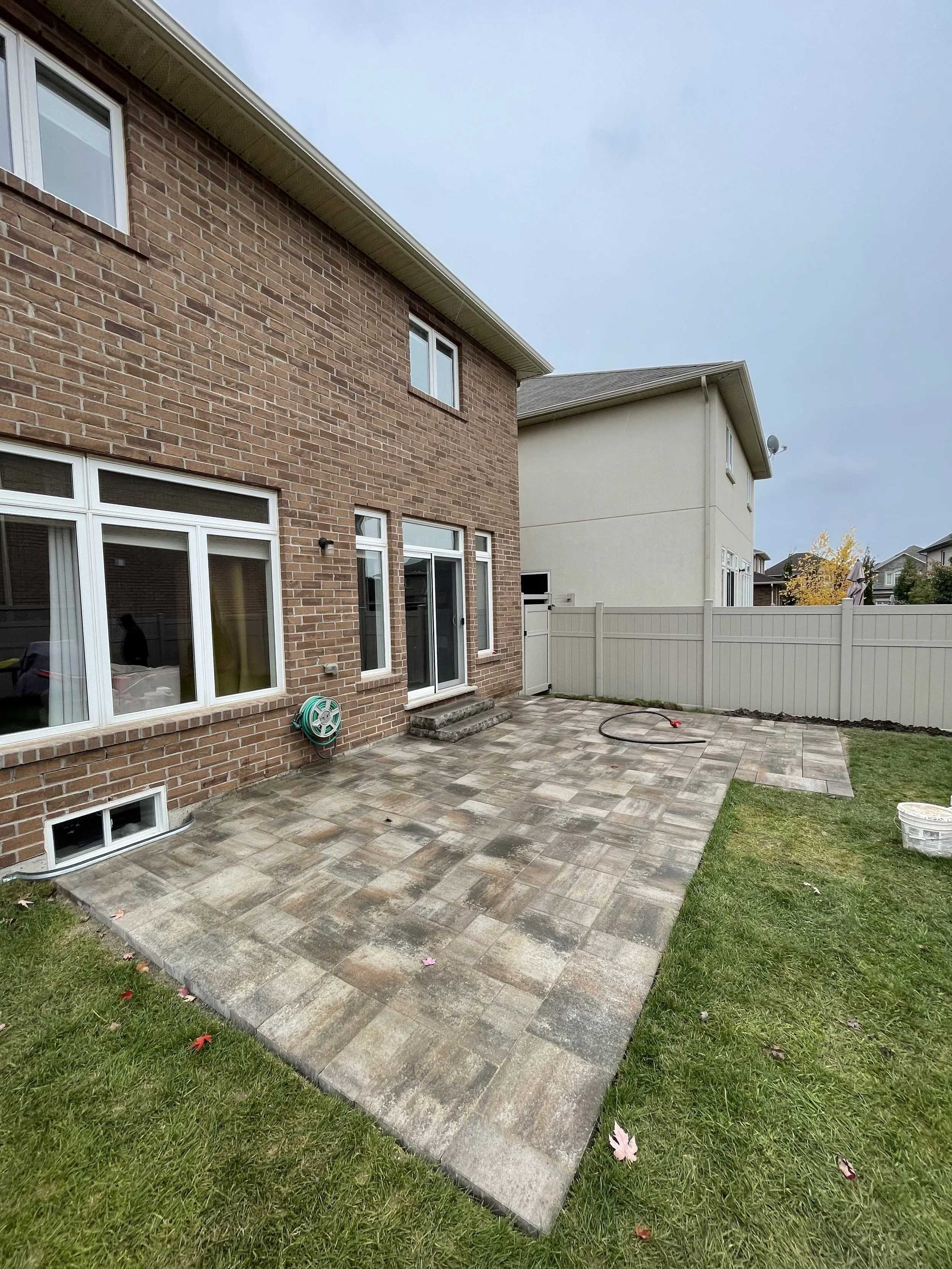 backyard patio with new stone pavers, a garden hose reel, and a gray fence, adjacent to a brick house with large windows and steps leading to the house entrance.