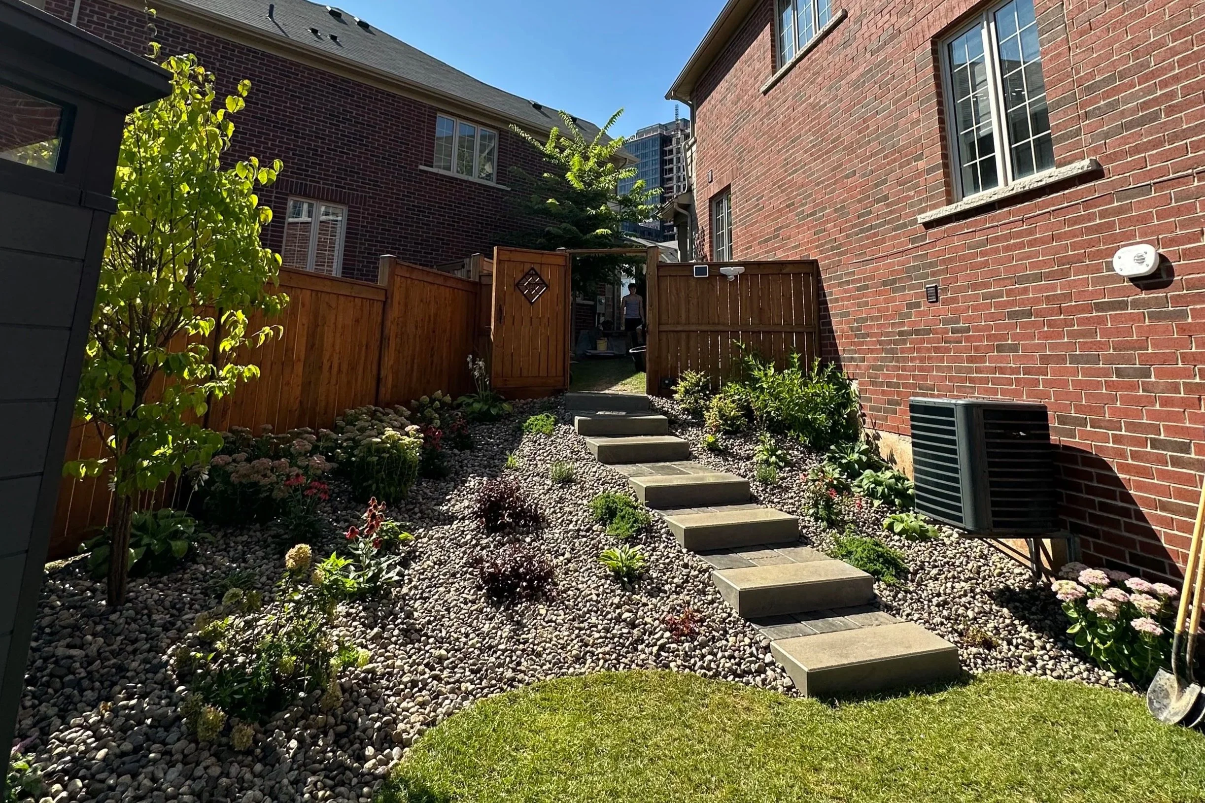 Backyard with a gravel garden, a pathway with square stepping stones, a wooden fence, and a brick house wall with windows, air conditioning unit, and garden plants.