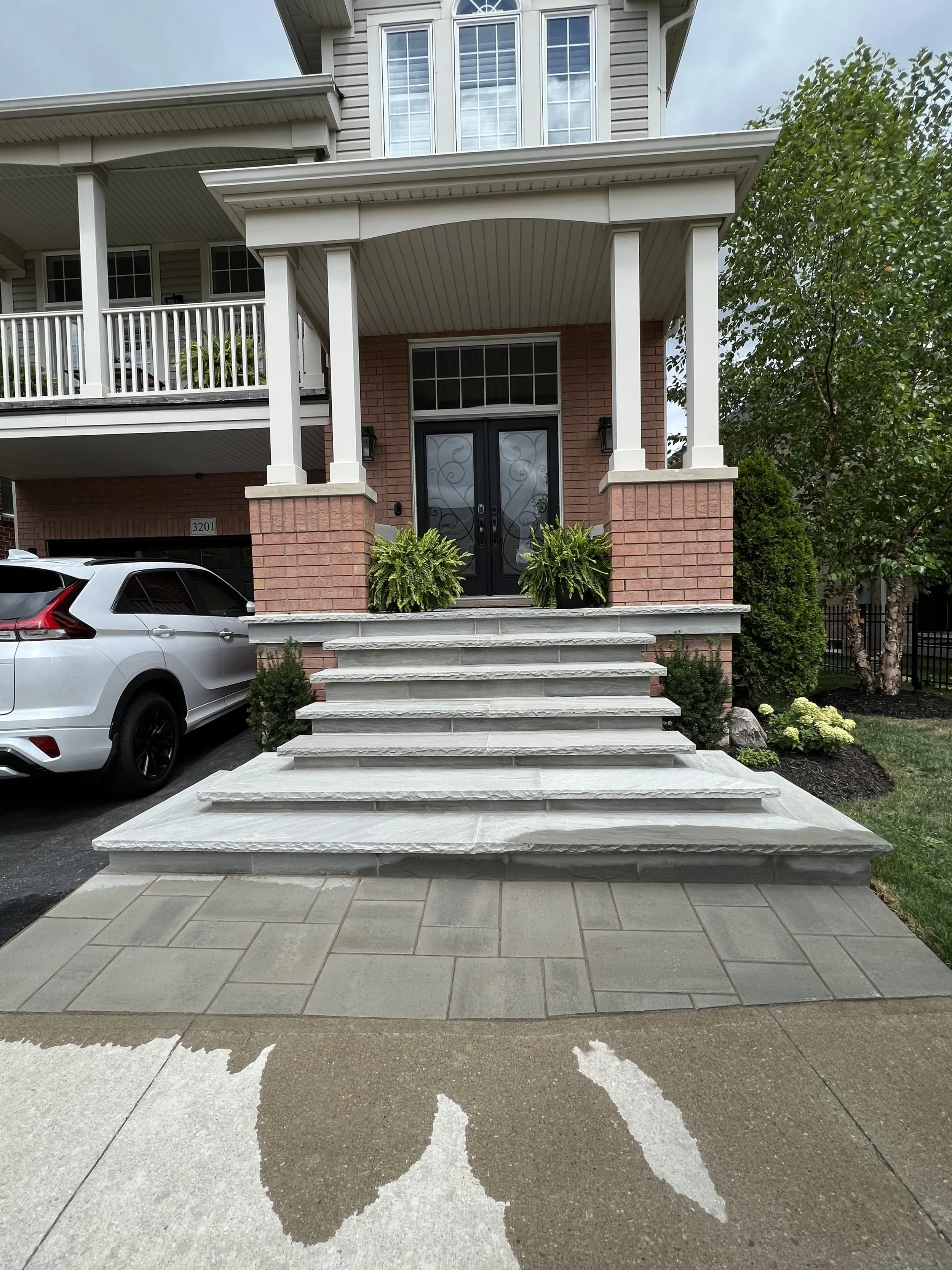The image shows the front entrance of a modern house with a brick and siding exterior, a set of wide steps leading up to black double doors, two potted plants on each side of the door, a white car parked to the left, and some greenery including trees