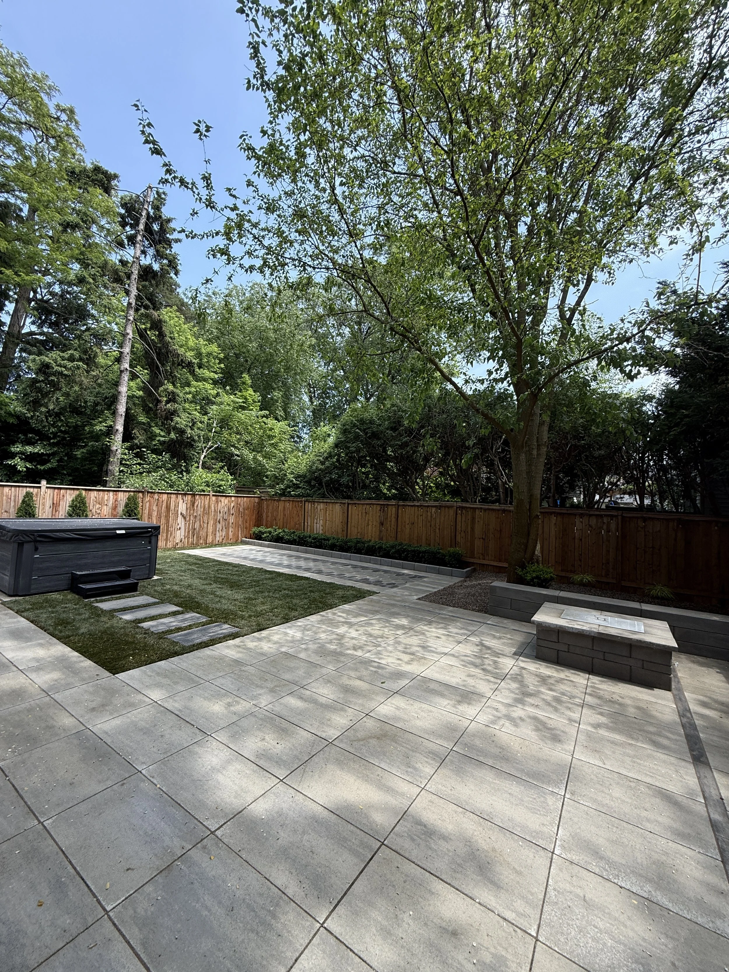 A backyard with a tiled patio, a hot tub, a grassy area, a paved walkway, a large tree, and a wooden fence, under a clear blue sky.