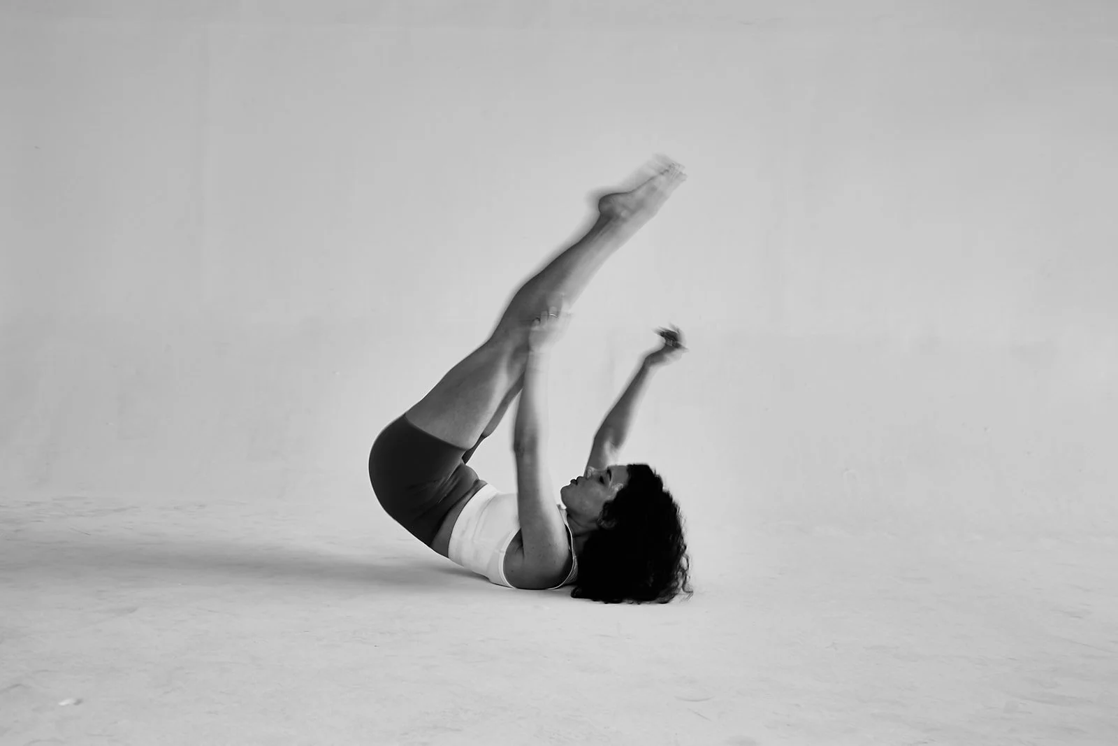 A woman performing a Pilates exercise on the floor, lying on her back with her legs extended upwards and her arms reaching towards her feet, in a minimalist room.