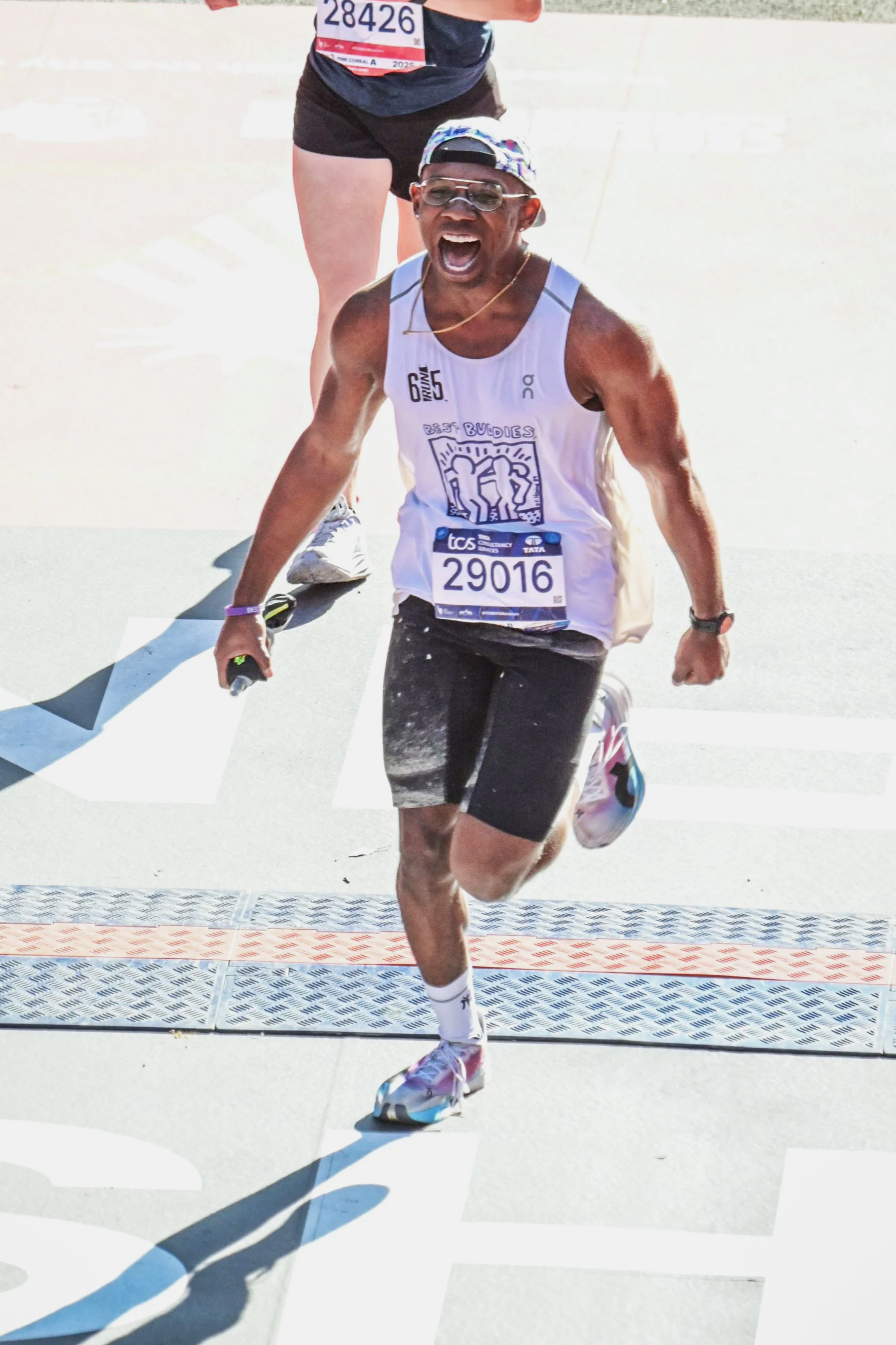 Male marathon runner crossing the finish line, wearing bib number 29016, shorts, tank top, sunglasses, and a cap, with a joyful expression.