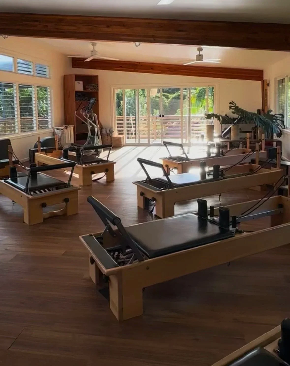 Pilates studio with multiple reformer machines arranged in a spacious room with natural sunlight coming through large windows and sliding glass doors, wooden ceiling accents, and a plant in the corner.