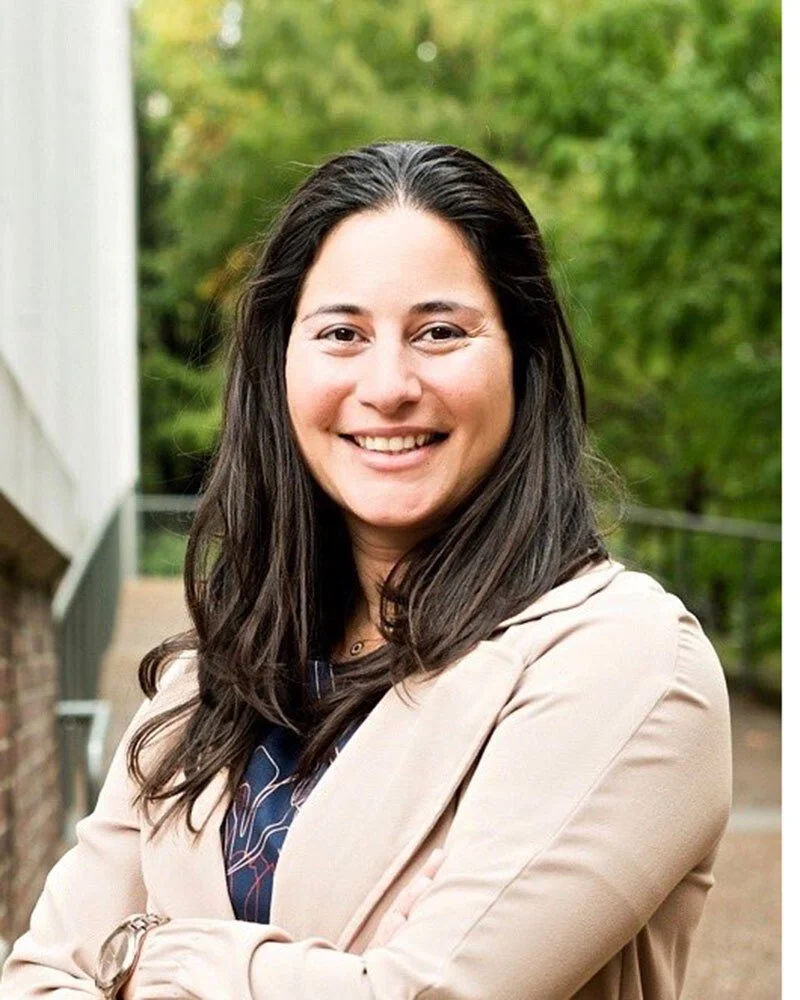 A woman with long dark hair smiling and crossing her arms, standing outdoors with green trees in the background.