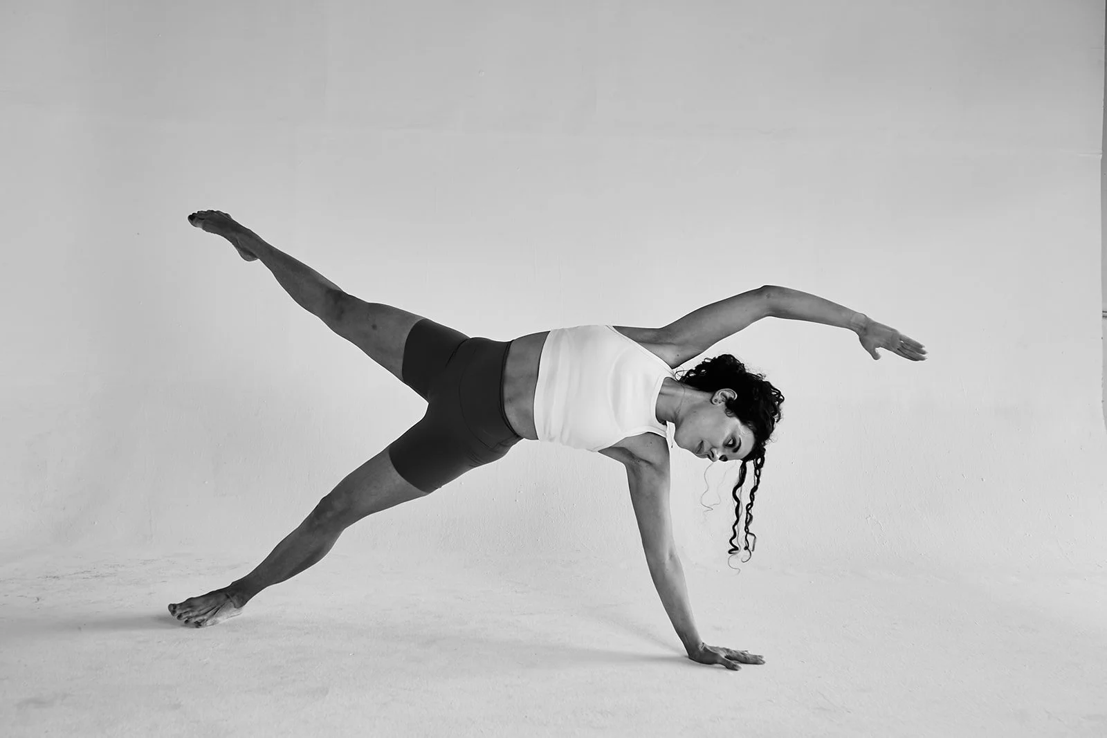 A woman performing a Pilates exercise with one arm extended down and the other arm extended upward, balancing on one hand and one foot, in a studio with a plain wall background.