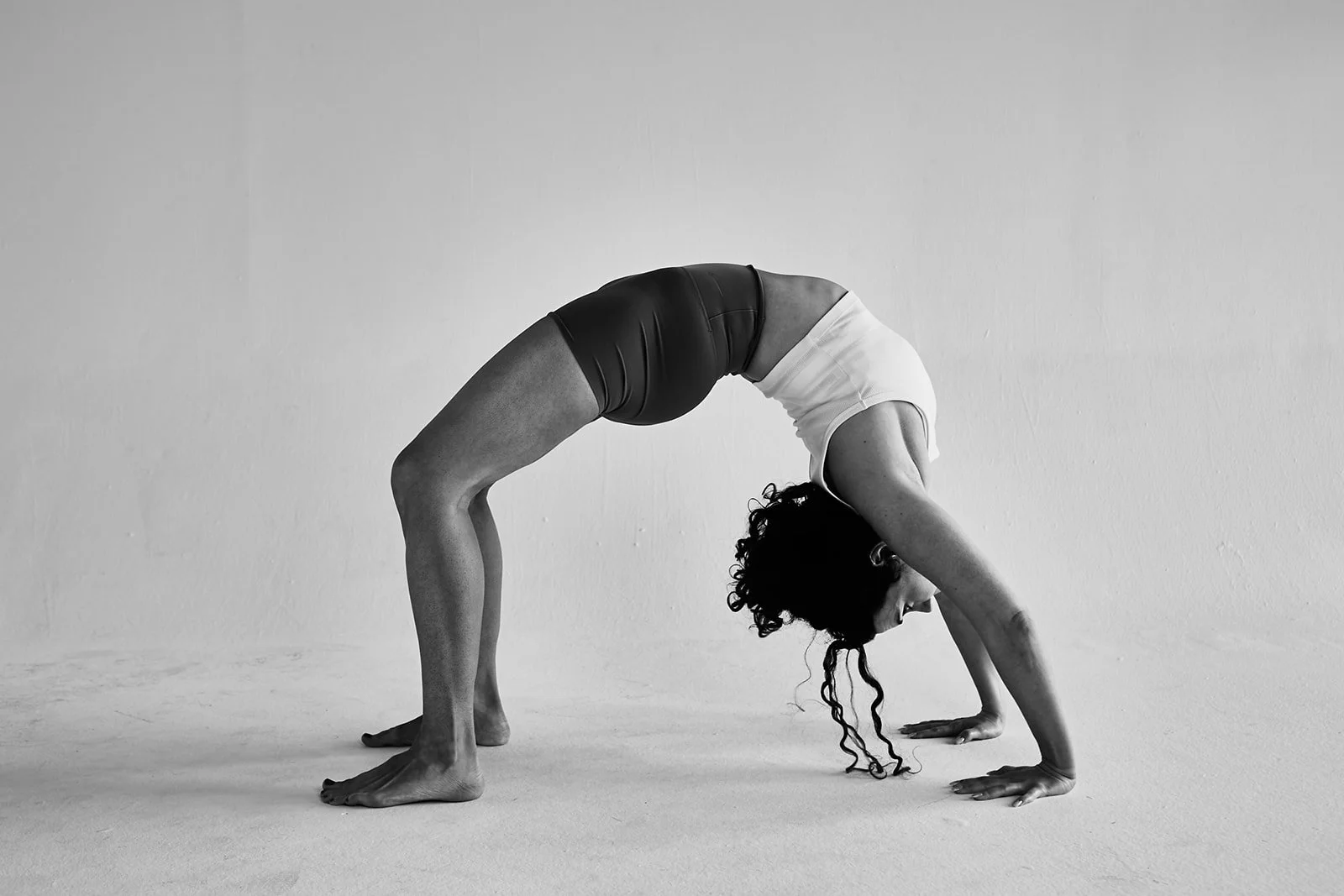A woman performing a backbend yoga pose on a plain floor with a blank wall background.