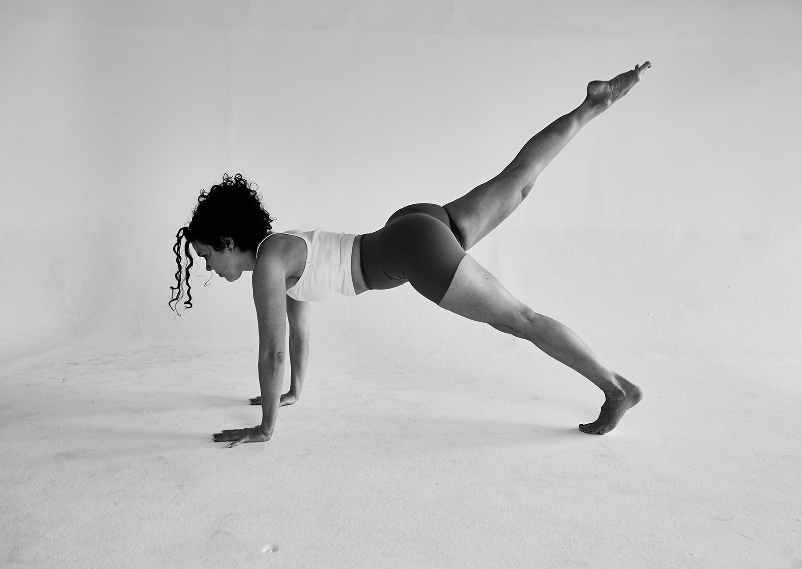 A woman in workout clothes performing a Pilates exercise on a smooth floor, with one leg extended upward and arms supporting her on the ground.