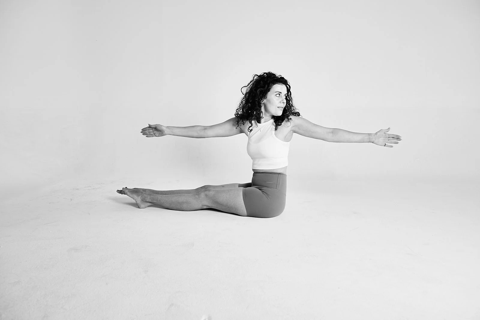 A woman practicing yoga indoors, sitting on the floor with her legs extended, arms outstretched to the sides, with a focused expression.