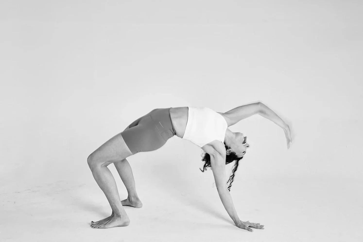 Black and white photo of a woman in a yoga pose on a plain white background. She is in a bridge pose with one arm reaching over her head and the other supporting her on the ground.