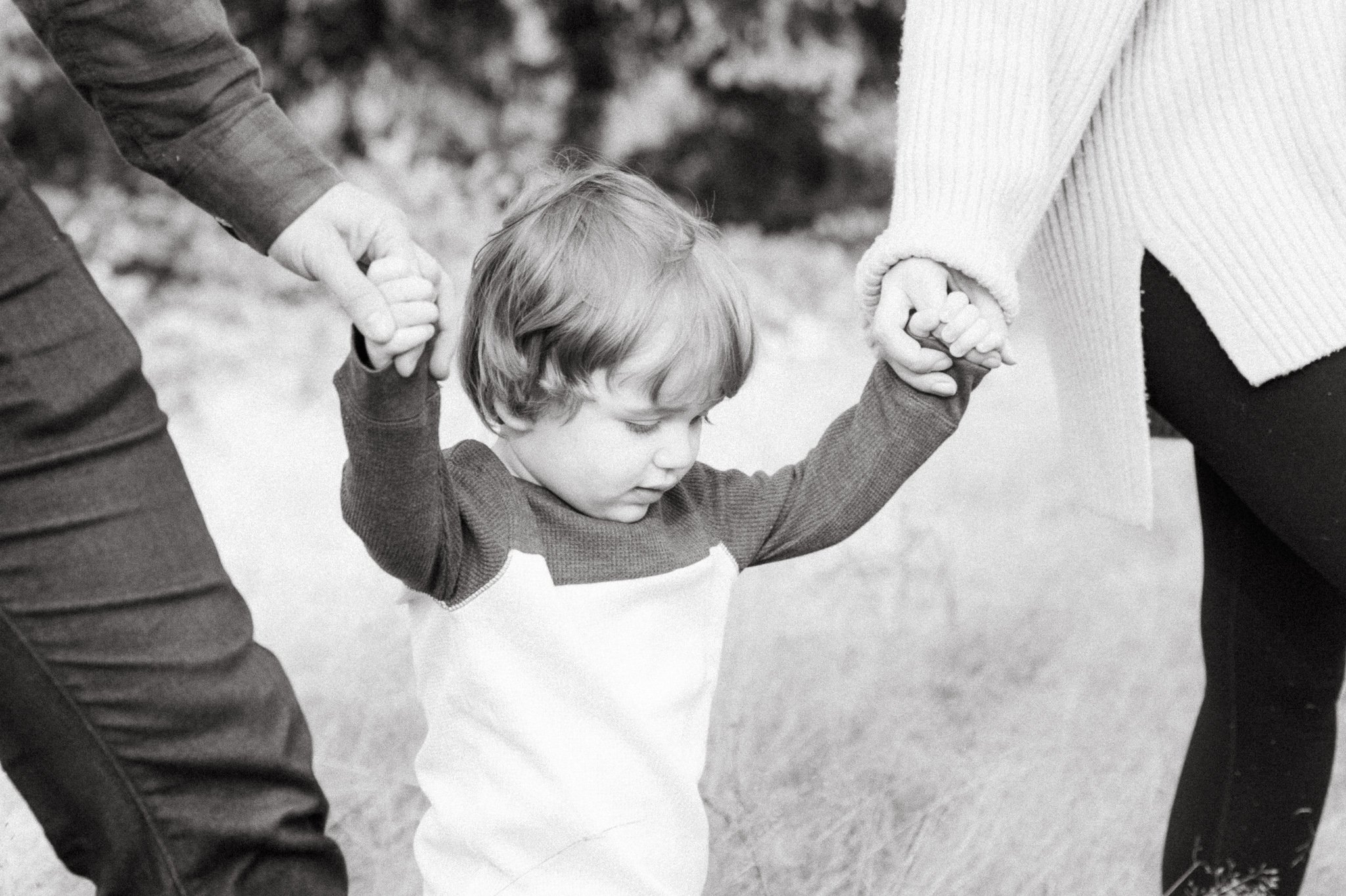 A young child holding hands with two adults outdoors, walking on grass.