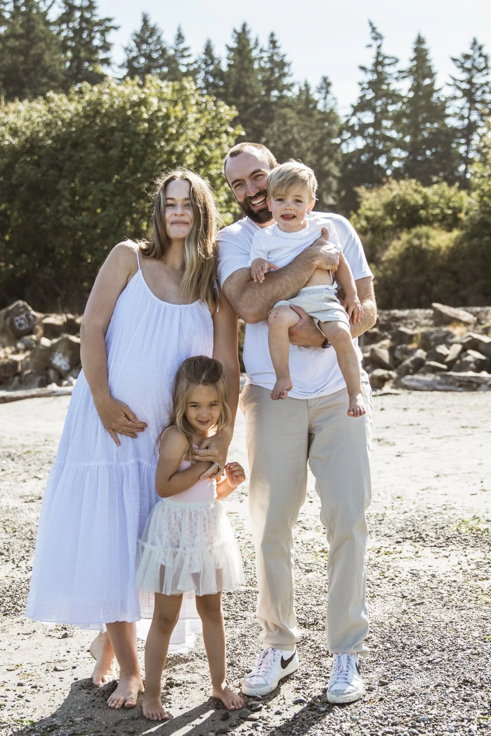 A family of five standing outdoors on a sandy beach with trees in the background, smiling and enjoying the sunny day.