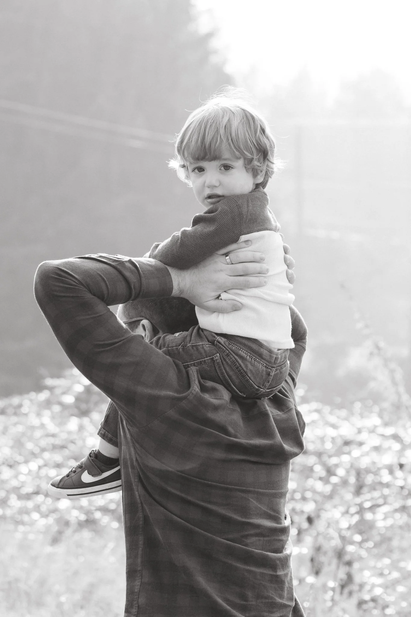 A child with light hair is sitting on an adult's shoulder, outdoors near a body of water, with trees and sunlight in the background.