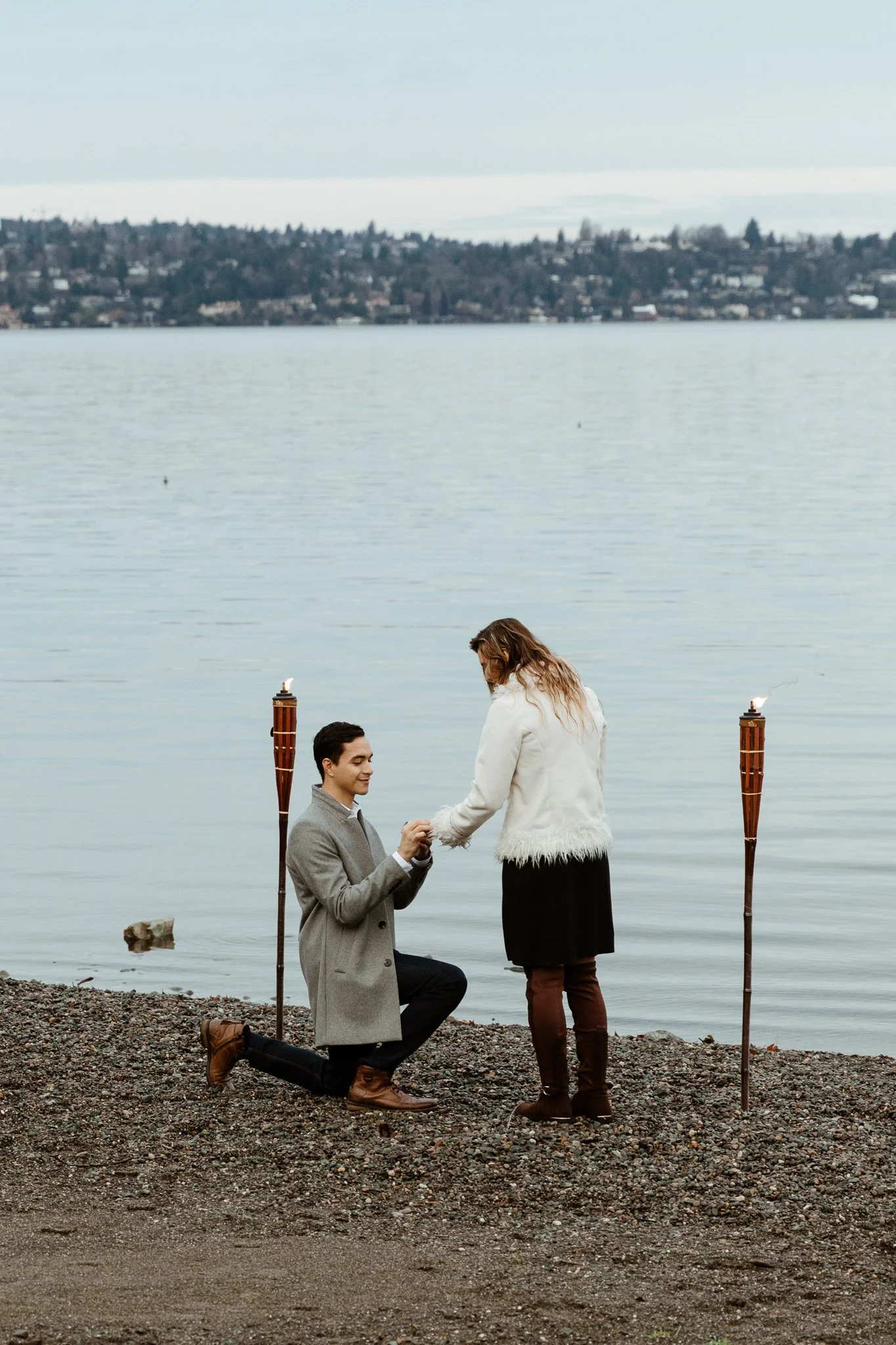 A man is kneeling on a pebbled shoreline, proposing marriage to a woman by holding her hands, near two tiki torches, with a large body of water and distant cityscape in the background.