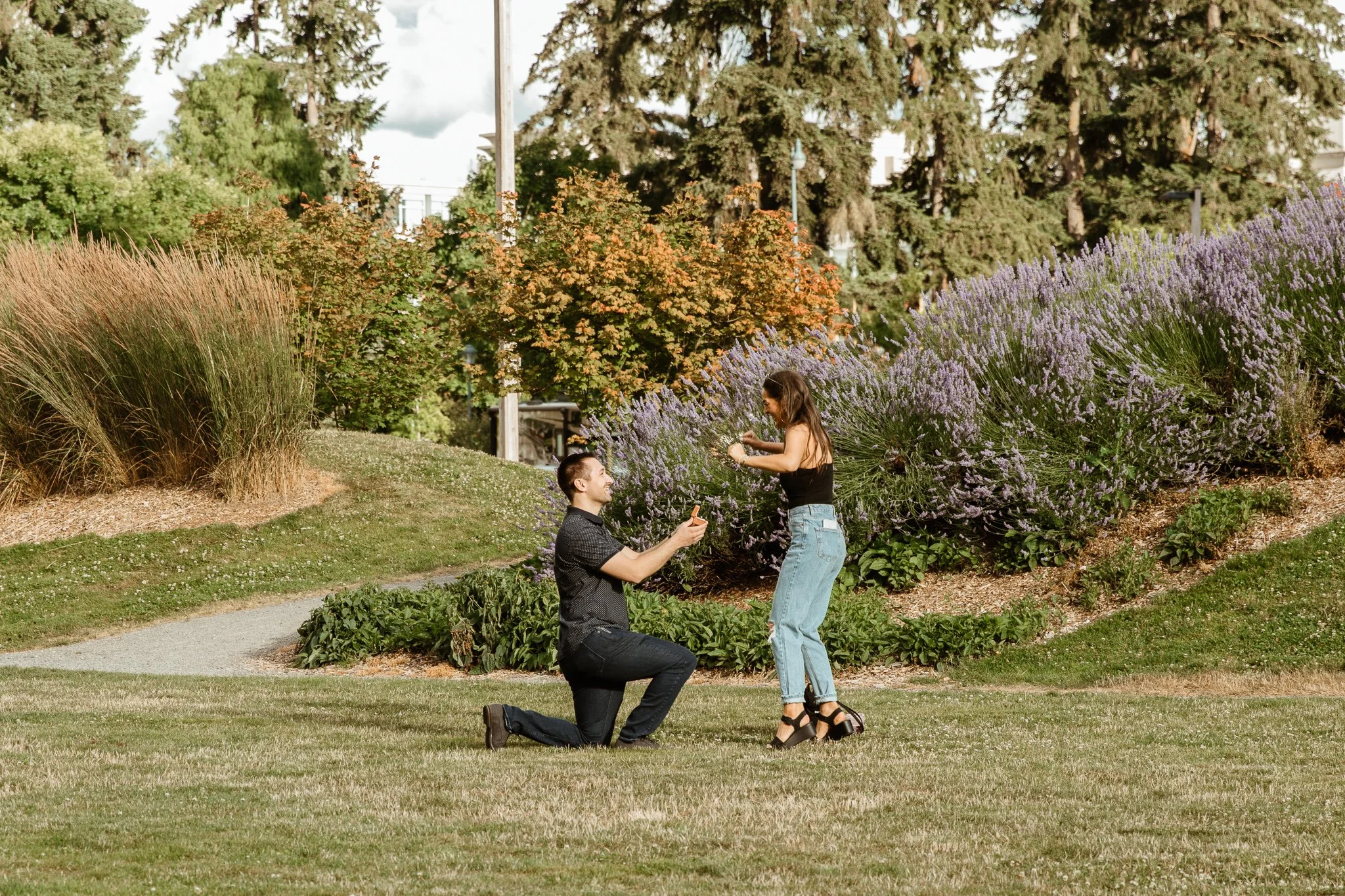A man proposing marriage to a woman in a park with colorful bushes and tall trees in the background.