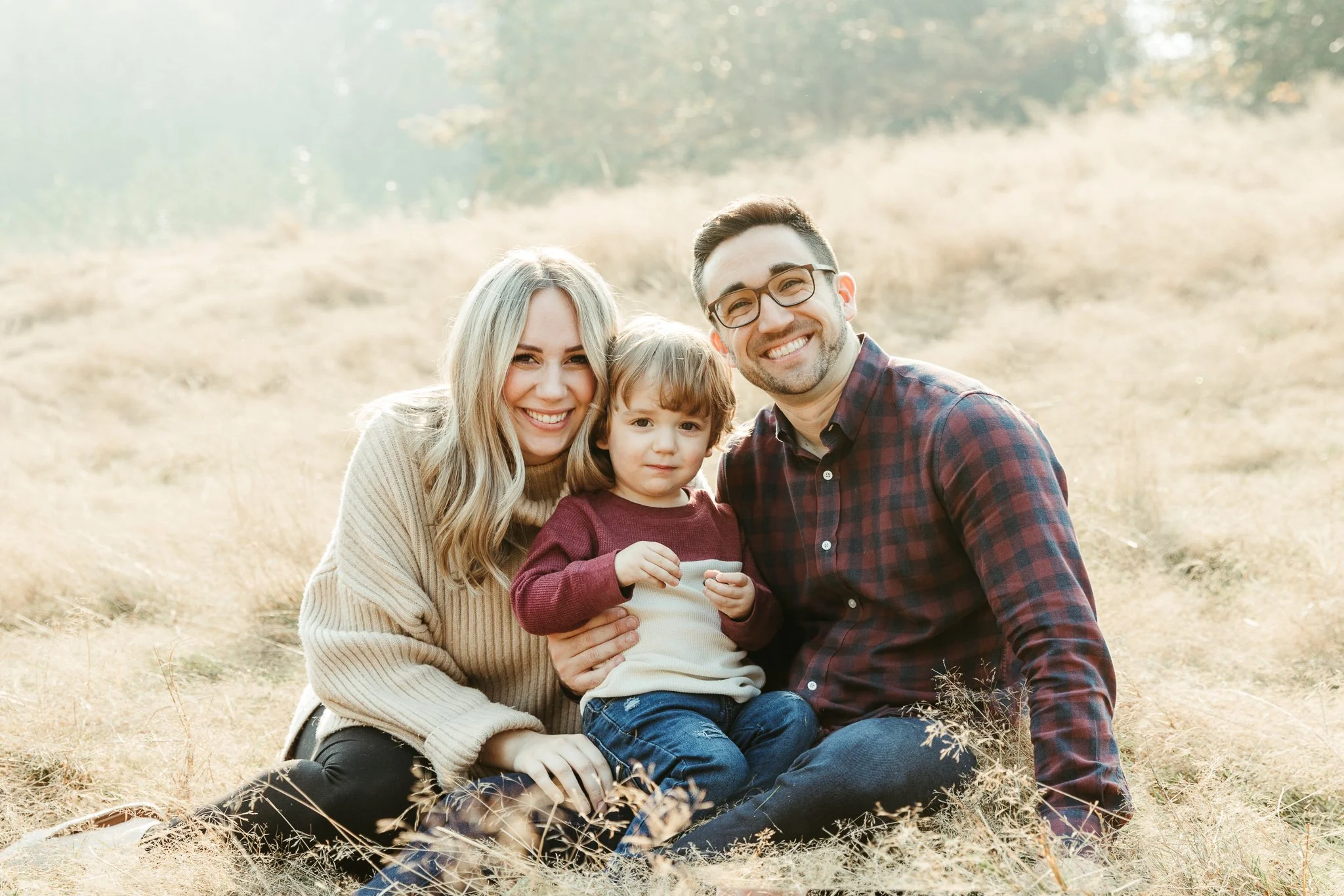 A happy family of three sitting on dry grassfield with trees in the background, smiling at the camera.