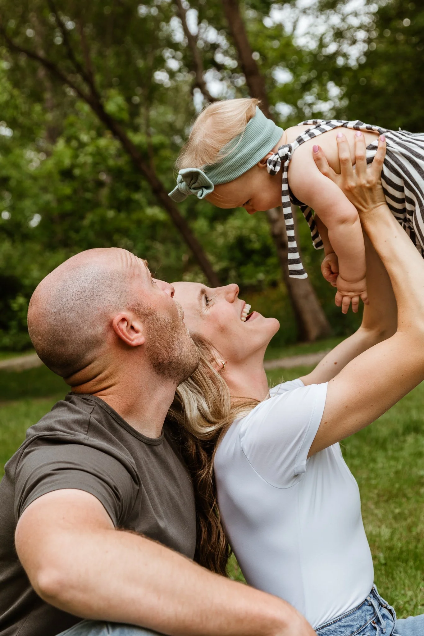 A family of three happily enjoying an outdoor moment, with the mother and father holding their baby girl up in the air. The background features green trees and grass, indicating they are in a park or garden.