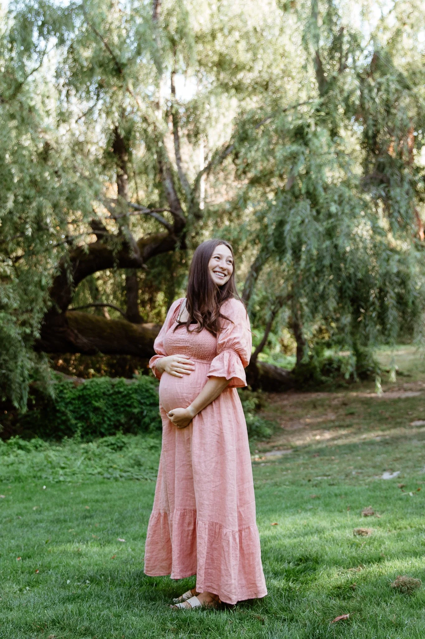 A pregnant woman in a pink dress stands on grass in a park with trees and sunlight in the background, smiling and looking to her right.