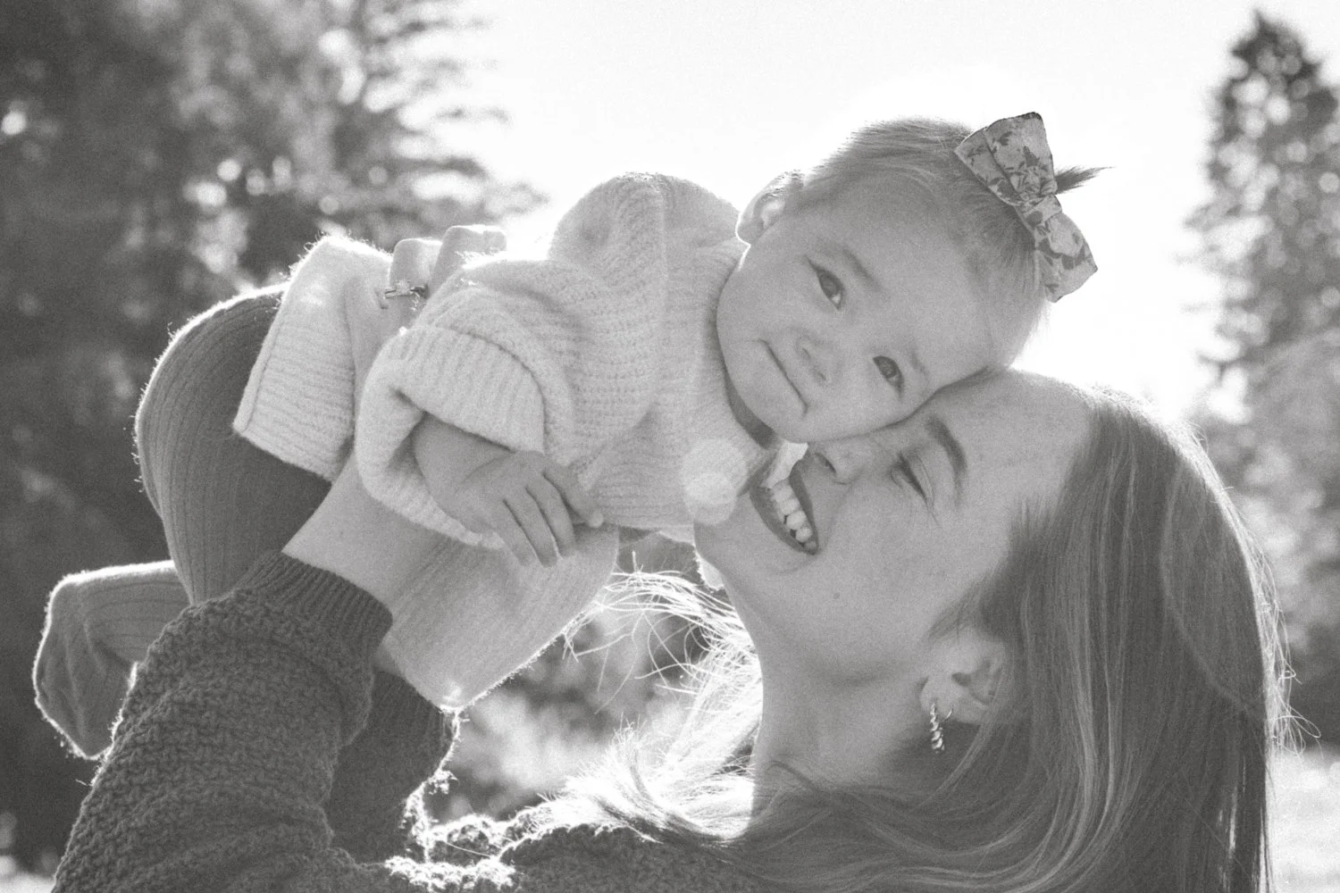 A woman holding a young girl close to her face, both smiling and enjoying a moment outdoors in sunlight, with trees in the background.