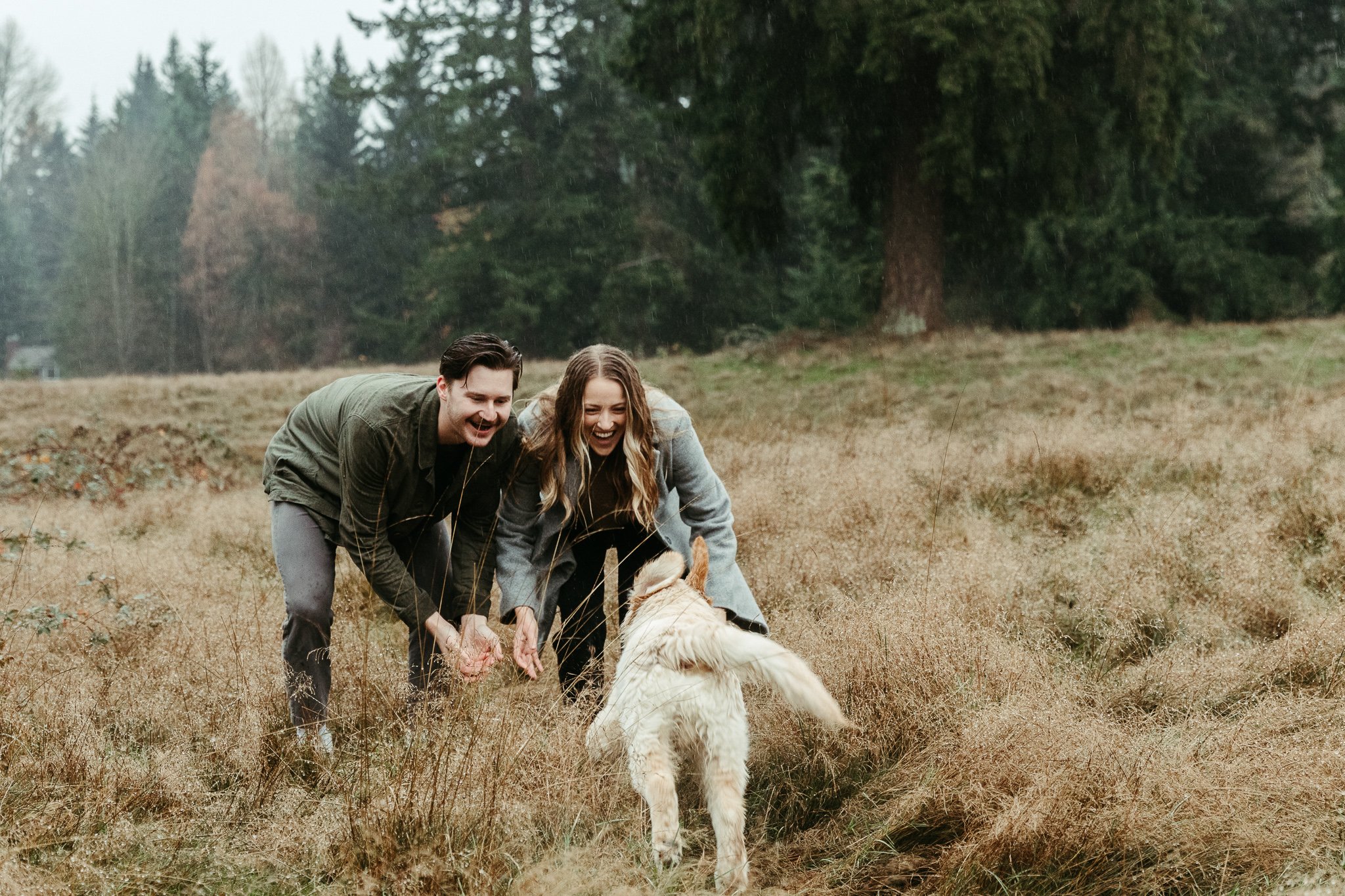 A happy couple playing with a dog in a grassy outdoor field with trees in the background.