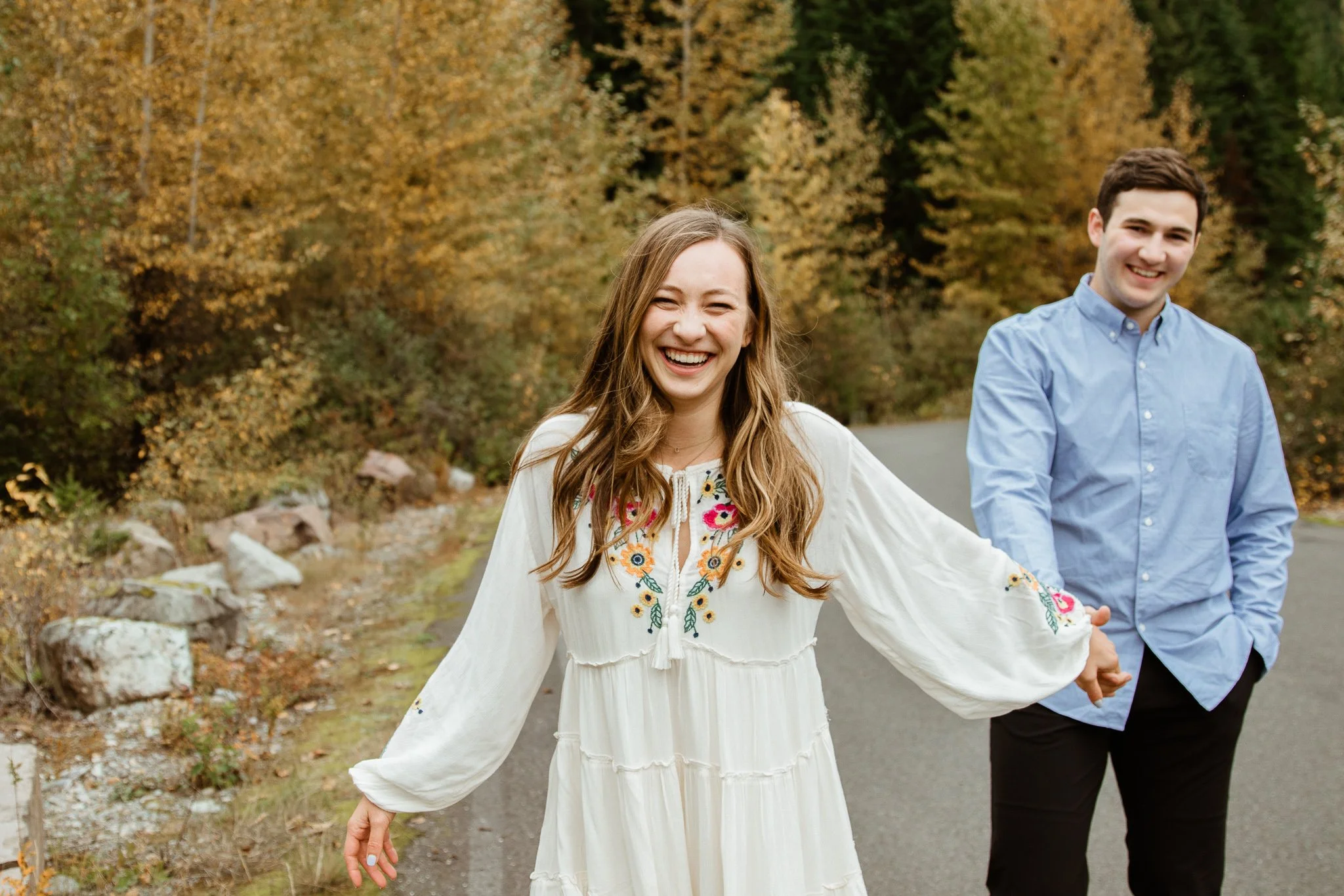 A young woman in a white embroidered dress smiling and holding hands with a young man in a light blue shirt, outdoors with autumn foliage in the background.