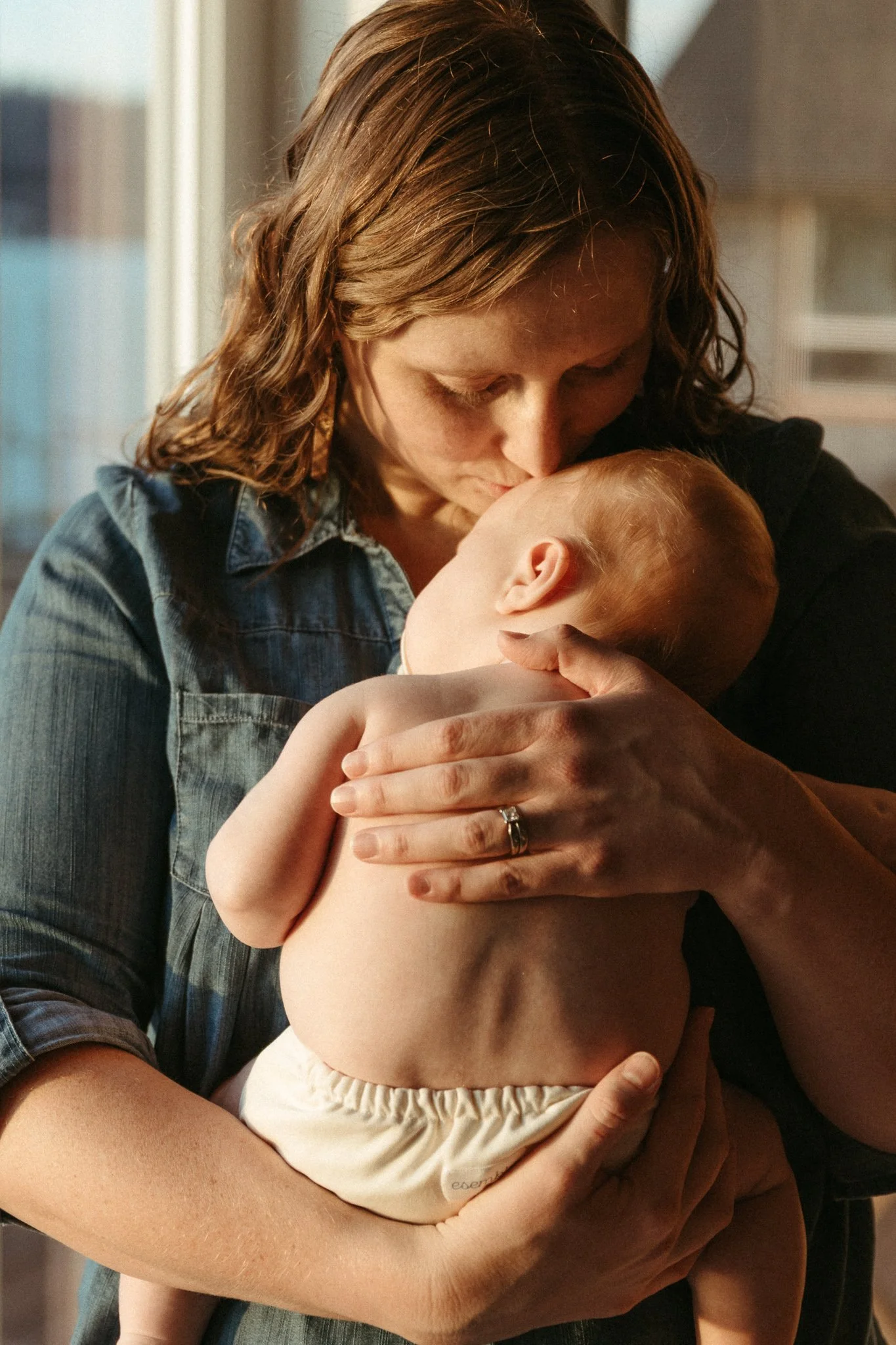 A woman with shoulder-length brown hair holding and kissing a small, shirtless baby in her arms, in a warmly lit room.