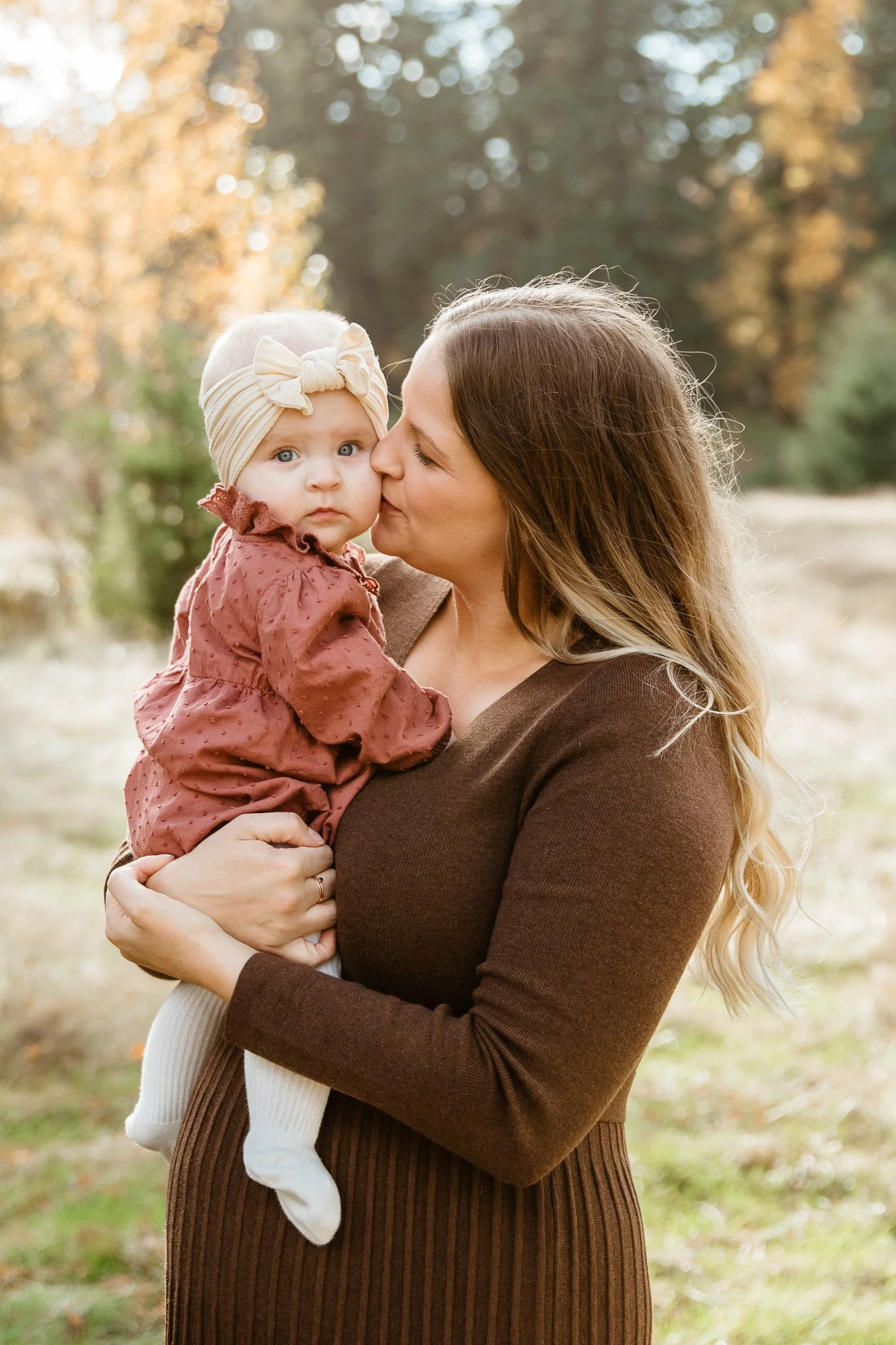 A woman holding a baby girl outdoors with trees and fall foliage in the background.