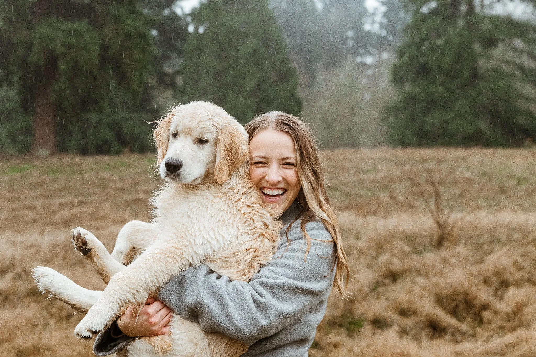 A woman smiling broadly while hugging a wet golden retriever puppy outdoors during rain, with trees and grass in the background.