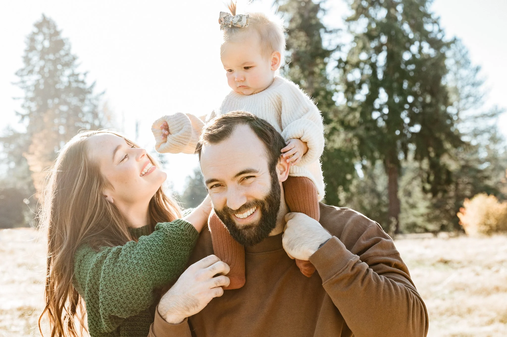A family of three outdoors, a man with a beard carrying a young girl on his shoulders, a woman smiling and touching the girl, all enjoying a sunny day in a park with trees in the background.