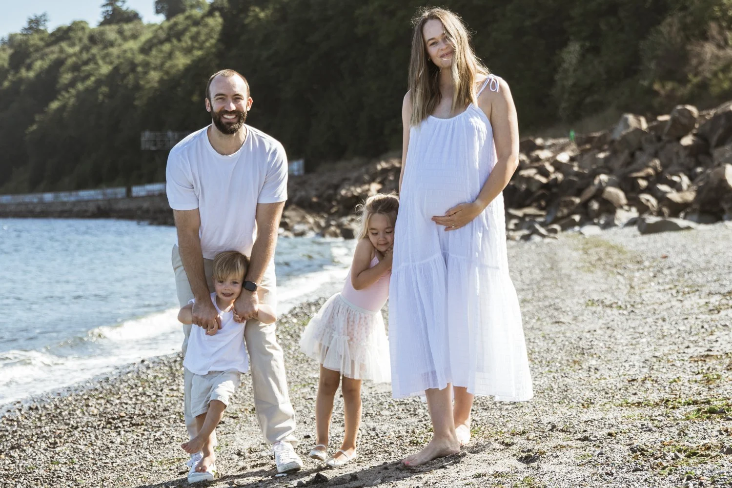 A family of four walking on a beach near water, with trees and rocks in the background. The pregnant mother is wearing a white dress, and the father and two children are dressed in light-colored clothing.