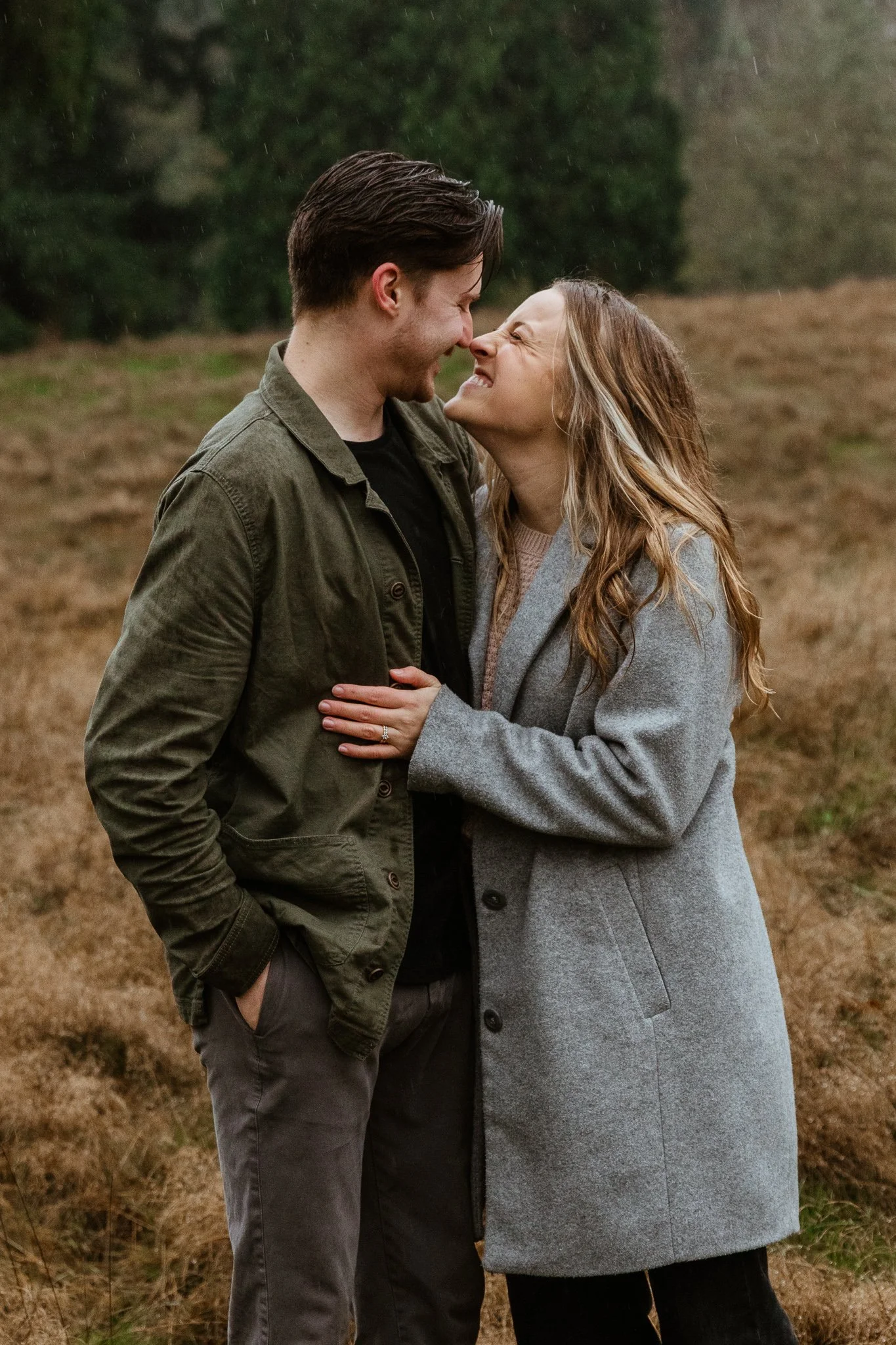 A couple standing close together outdoors, smiling and touching foreheads on a rainy day with a grassy field and trees in the background.