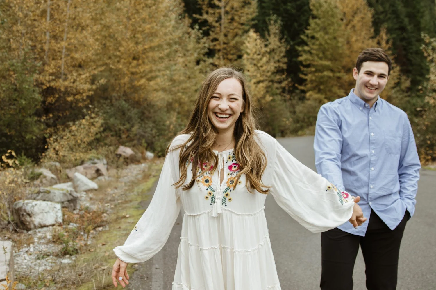 A smiling woman in a white embroidered dress holding hands with a smiling man in a blue shirt, standing on a fall-colored outdoor road.
